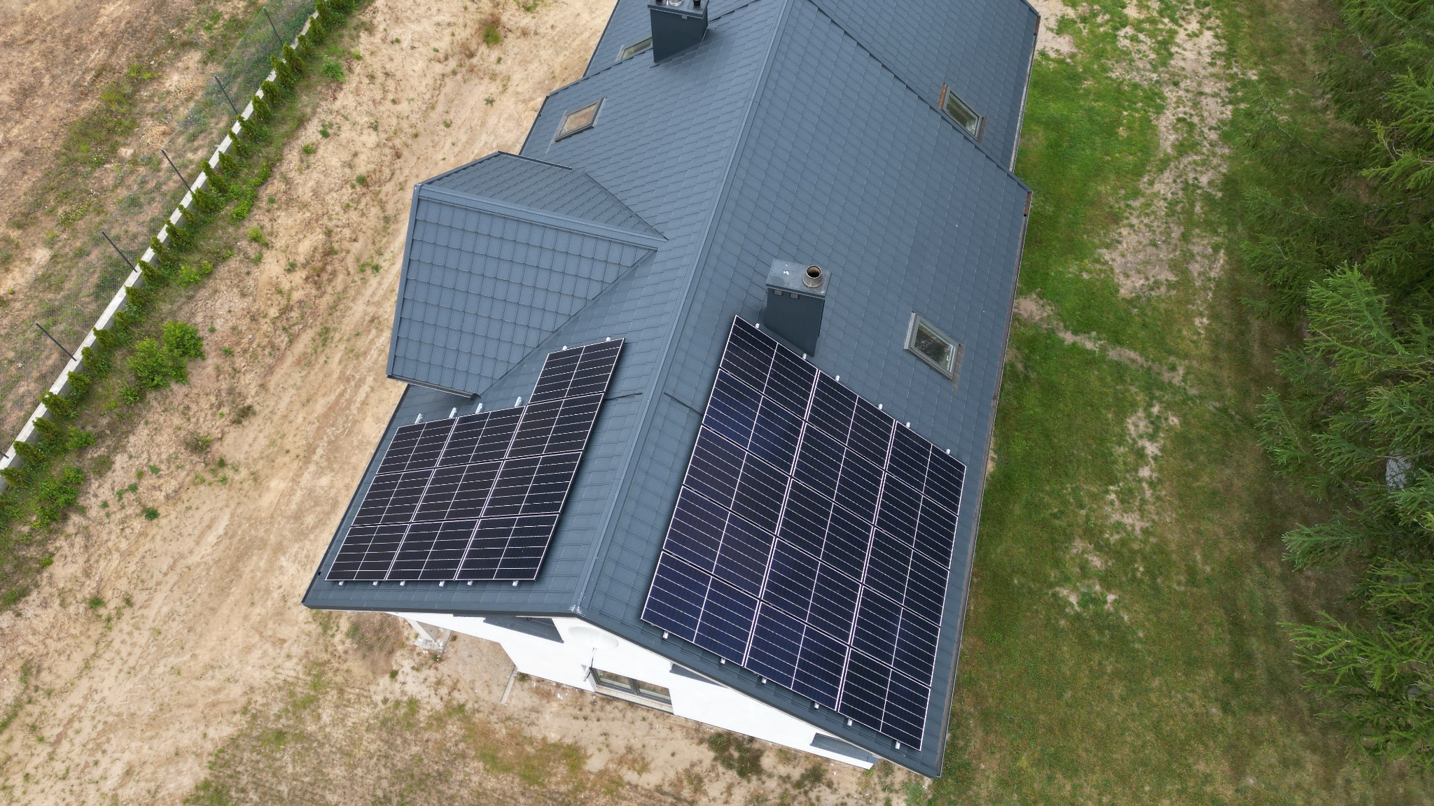Widok z góry na dom z szarym dachem i dwoma zestawami paneli słonecznych, otoczony piaszczystym terenem i zielenią.