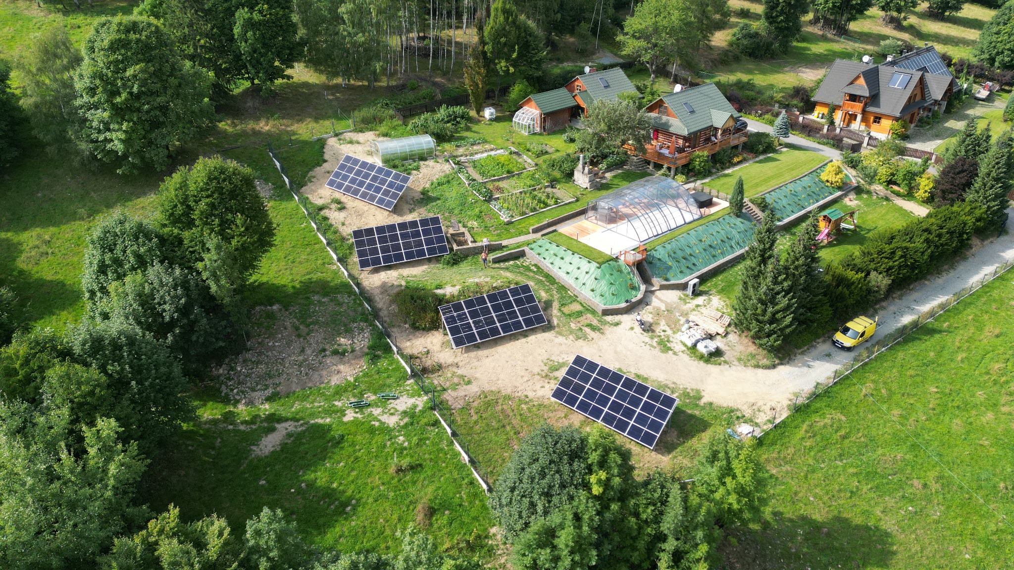 Widok z lotu ptaka na rozległą posesję z domami w stylu góralskim i kompleksem szklarni, na której zainstalowano panele słoneczne; żółty samochód dostawczy zaparkowany obok ogrodzenia.