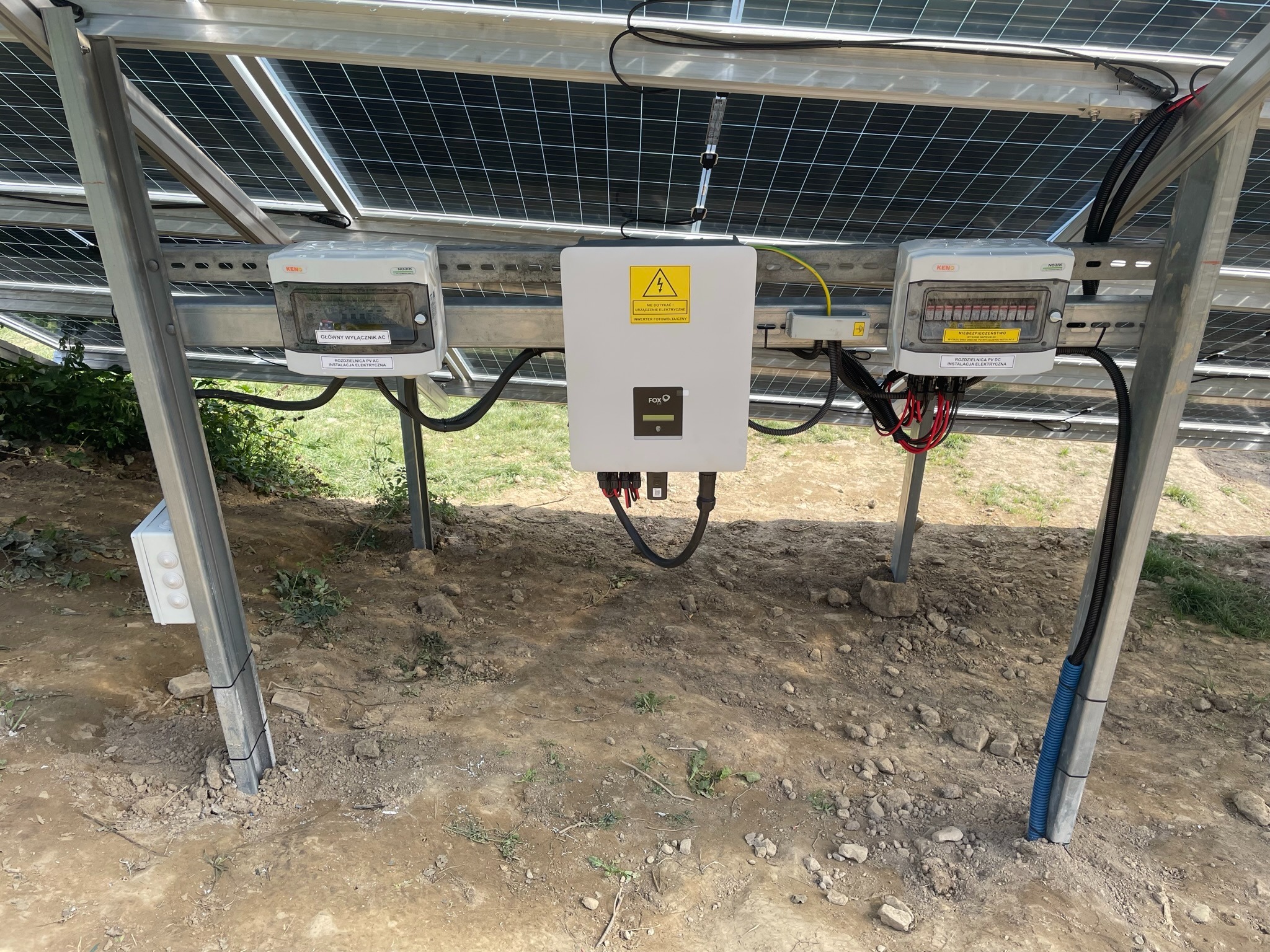 Instalacja paneli słonecznych: widok na inwerter FOX O, dwa rozdzielnice PV AC Noark oraz konstrukcję wsporczą paneli na gruncie z widocznymi przewodami i zabezpieczeniami elektrycznymi.