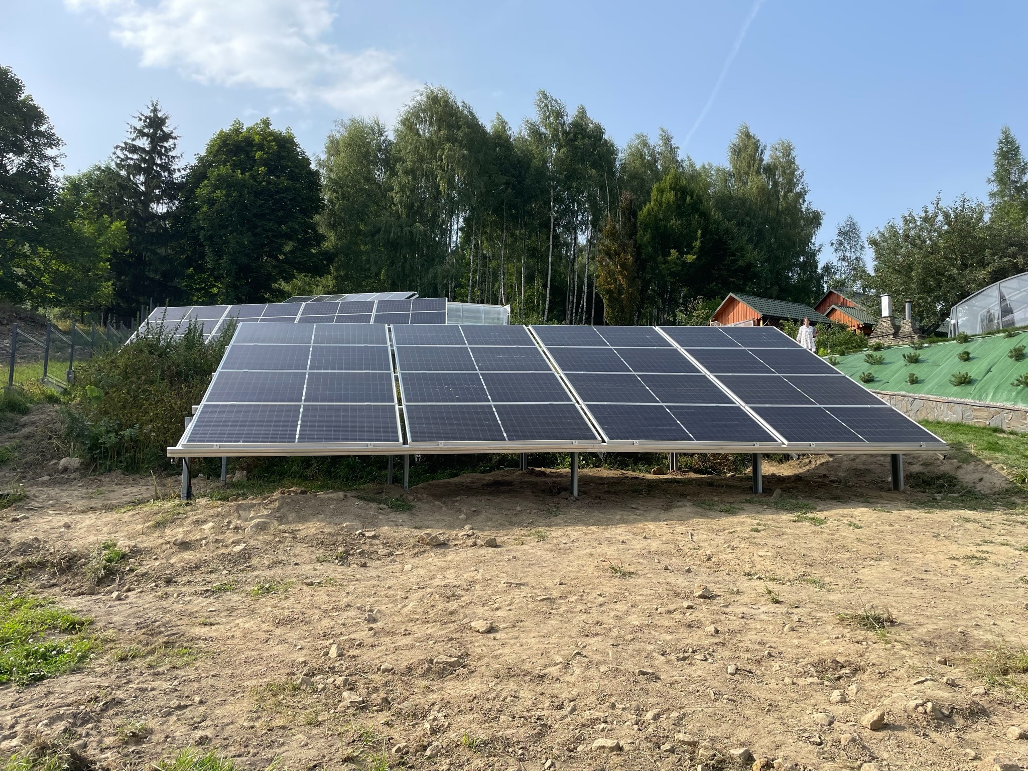 Instalacja fotowoltaiczna na metalowej konstrukcji, panele ustawione rzędami na nierównym, piaszczystym terenie, w tle drzewa i budynki.