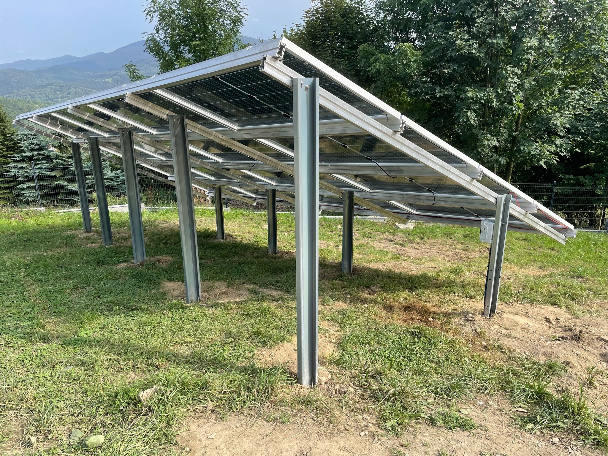 Instalacja paneli słonecznych na metalowej konstrukcji w Krze, widok od dołu z perspektywy trawnika, góry i drzewa w tle.