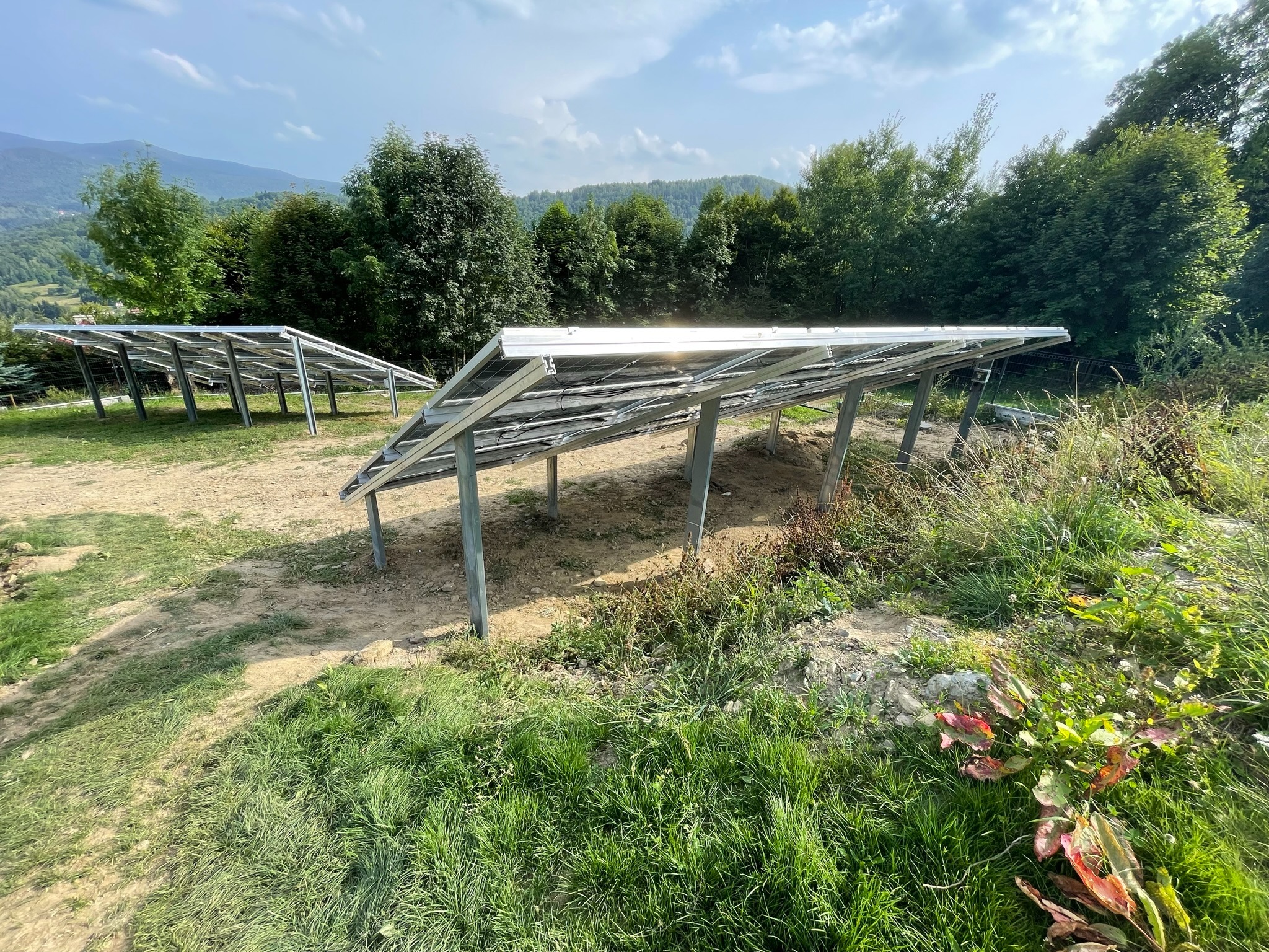 Metalowa konstrukcja pod panele słoneczne w trakcie montażu, widoczne metalowe słupki i ramy, w tle zieleń drzew i wzgórza.