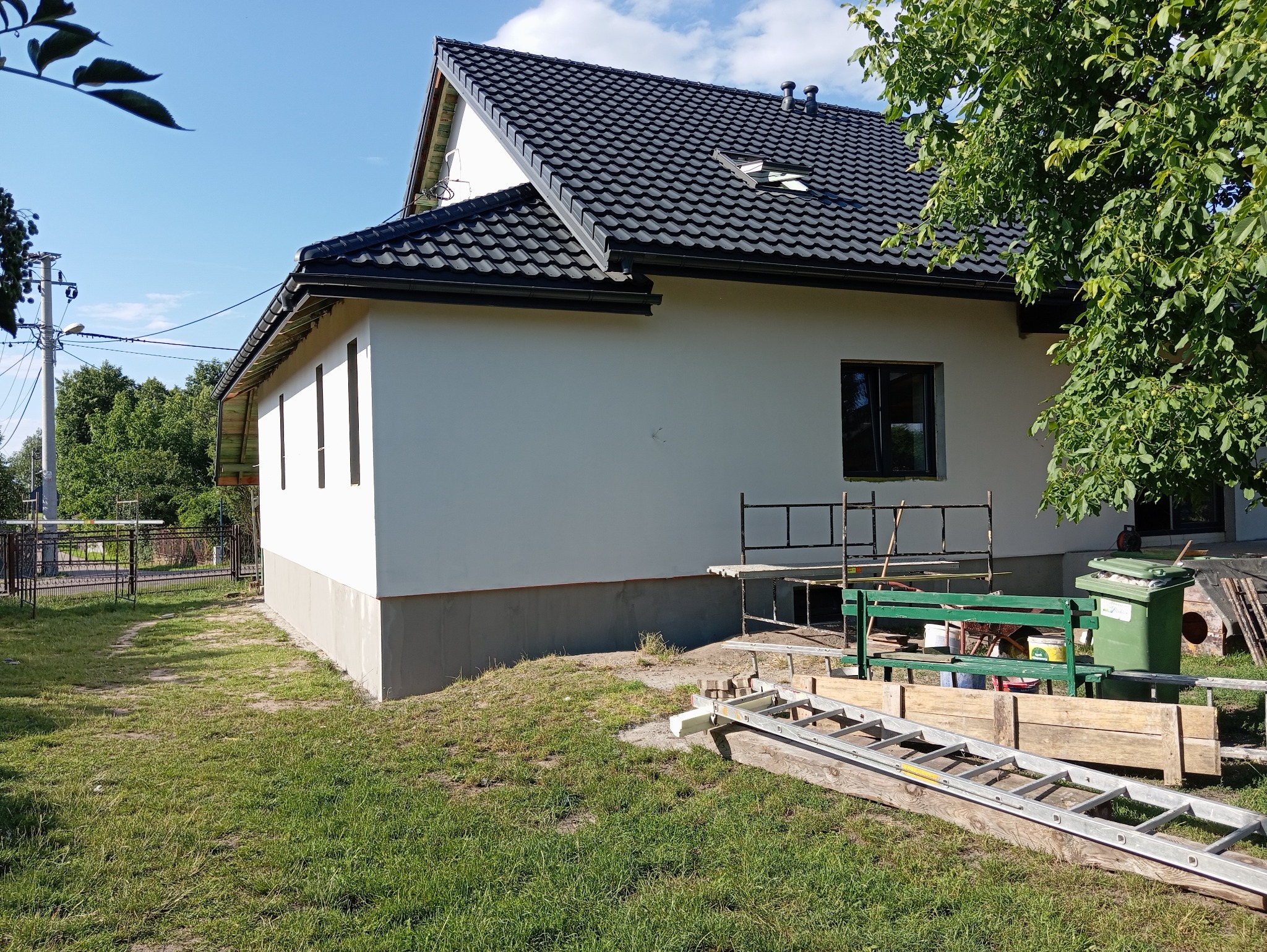 Ocieplony dom jednorodzinny z czarną dachówką, widok z boku na elewację po szpachlowaniu, z rusztowaniem, drabiną i narzędziami na trawniku.
