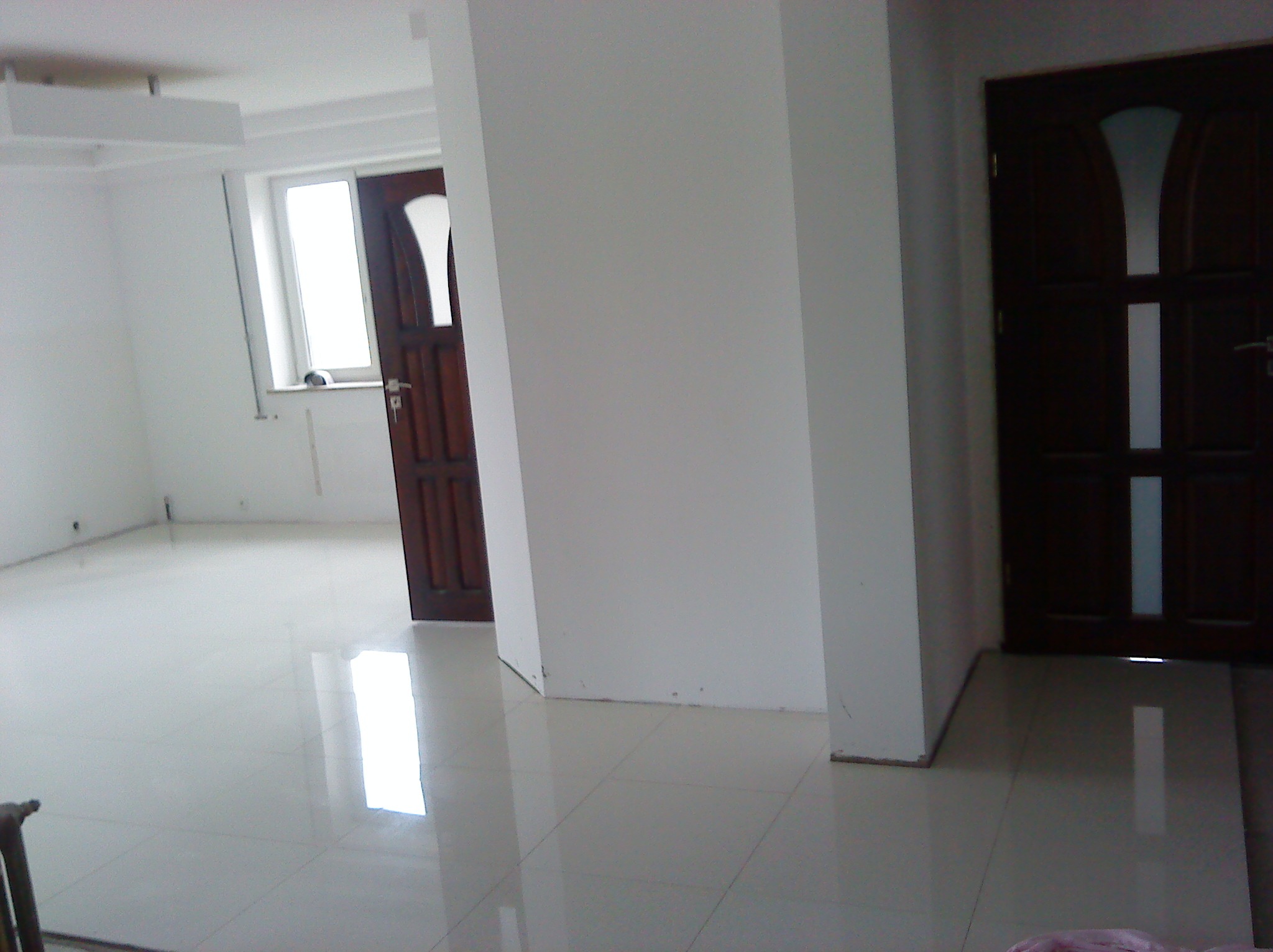 Jasne, przestronne wnętrze z nową, błyszczącą, kremową podłogą z płytek, widoczne dwie drewniane drzwi z przeszkleniami, ściany świeżo pomalowane na biało, w tle okno z widocznym fragmentem parapetu.