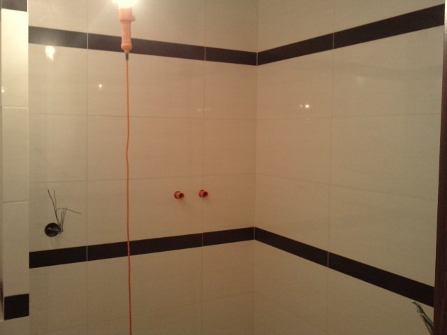 Ściana łazienki wyłożona jasnymi płytkami z ciemnobrązowym pasem dekoracyjnym, widoczne wystające kable i rurki, oświetlona wiszącą żarówką na kablu.