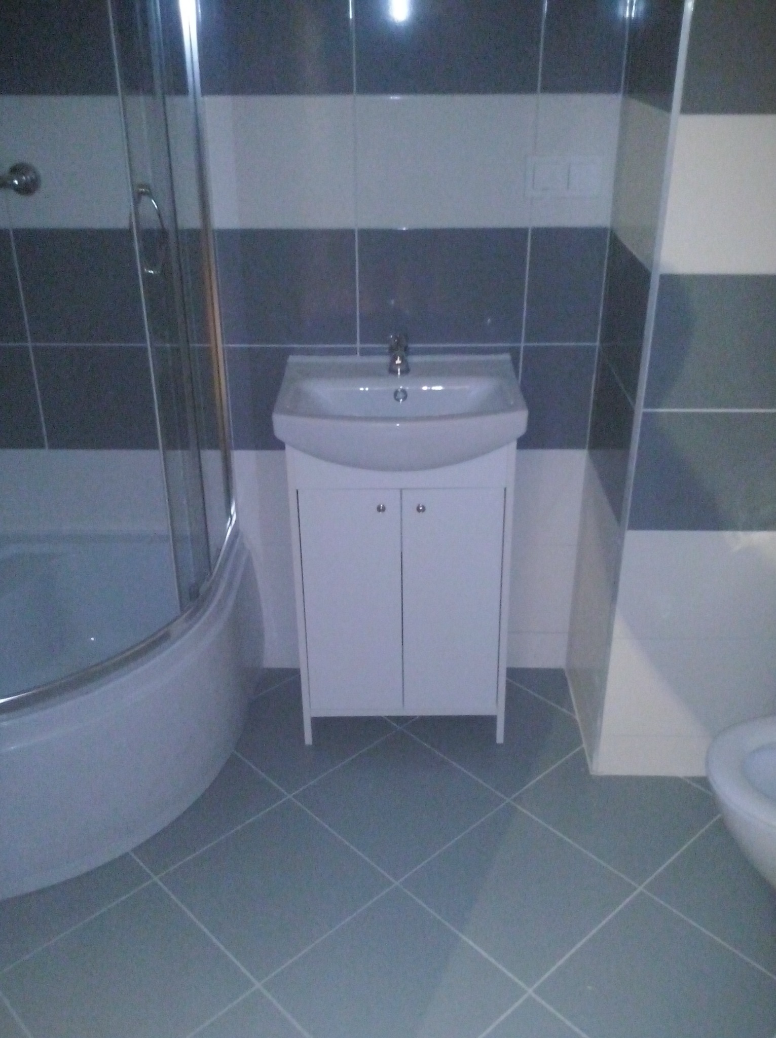 Nowoczesna łazienka z szaro-białymi płytkami w pasy, kabiną prysznicową z zaokrąglonym szkłem i białą szafką pod umywalką z dwoma drzwiczkami, wykończona szarą podłogą w romby.