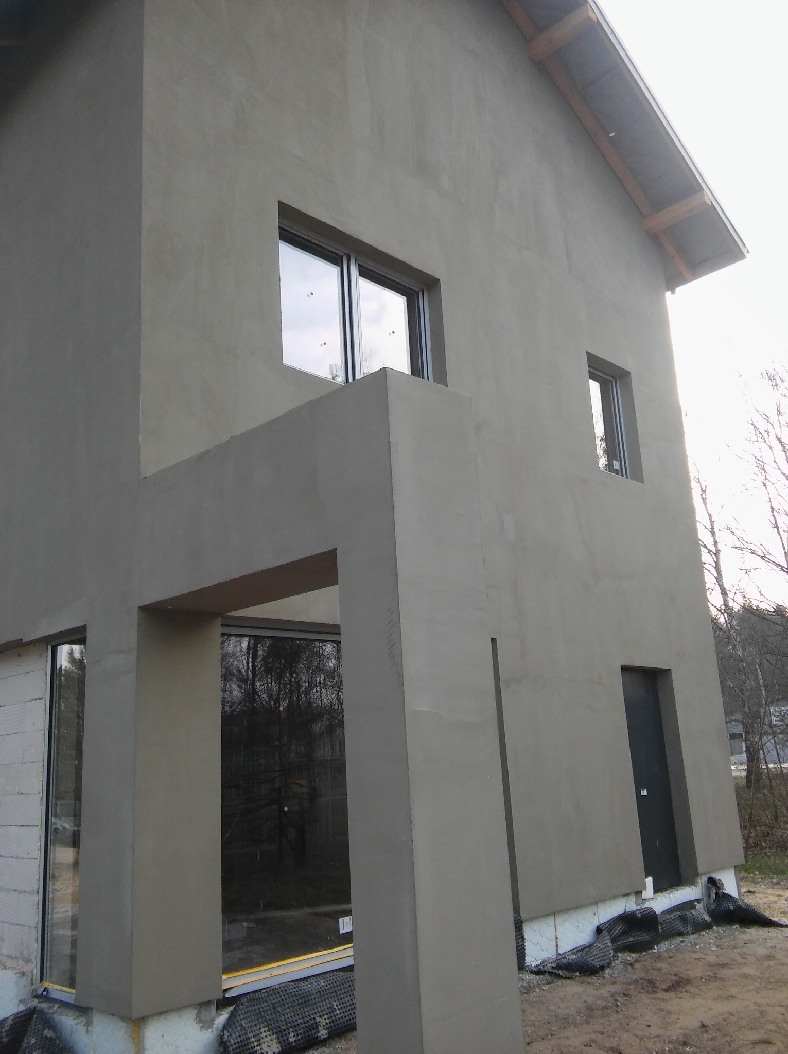Surowa elewacja domu jednorodzinnego w trakcie prac wykończeniowych, widoczne okna, drzwi wejściowe i fragment ocieplenia fundamentów.