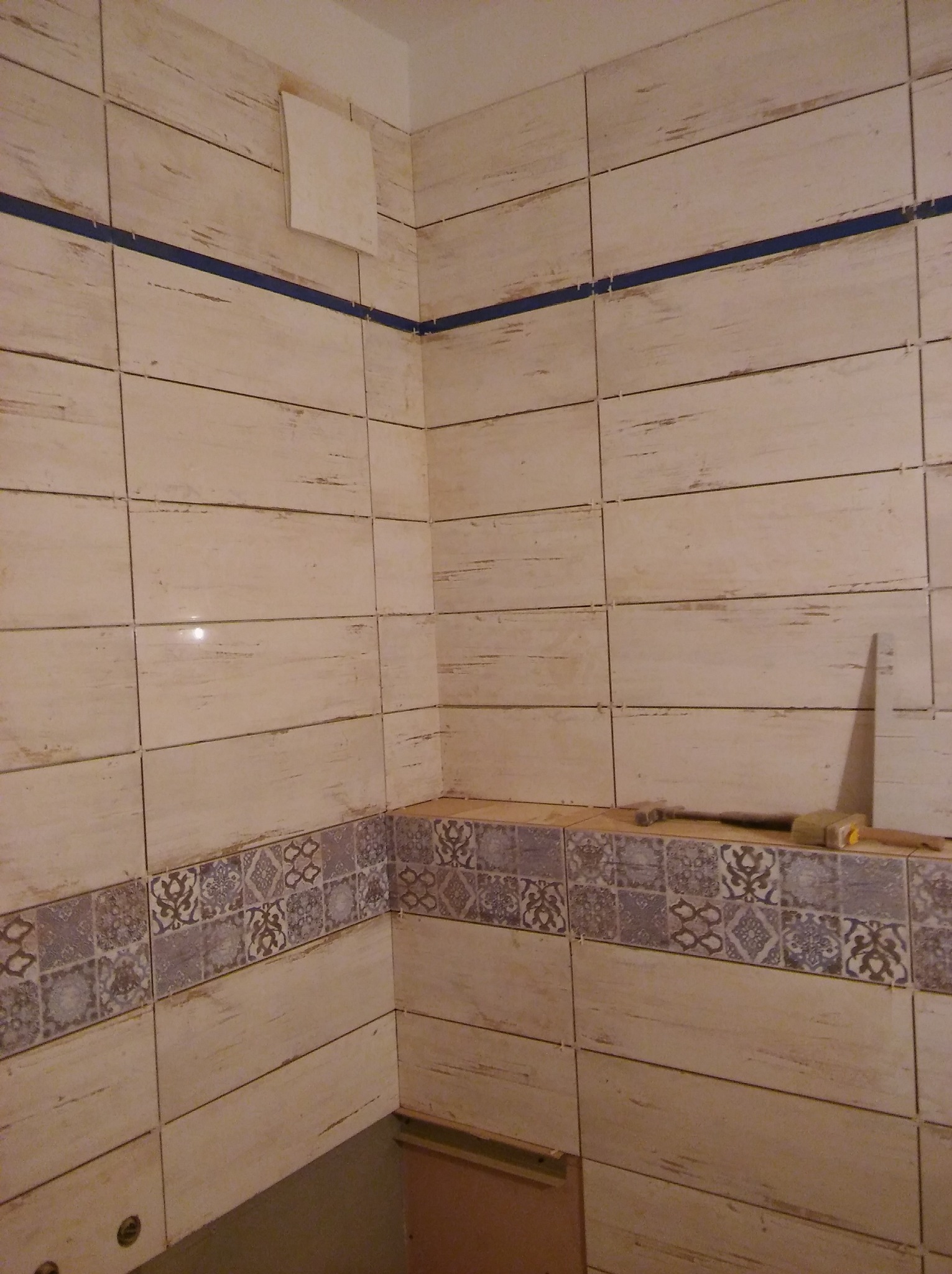 Ściana w łazience wyłożona beżowymi płytkami imitującymi drewno, z dekoracyjnym pasem płytek z motywami roślinnymi, narzędzia glazurnicze na półce, widoczny fragment wentylacji.