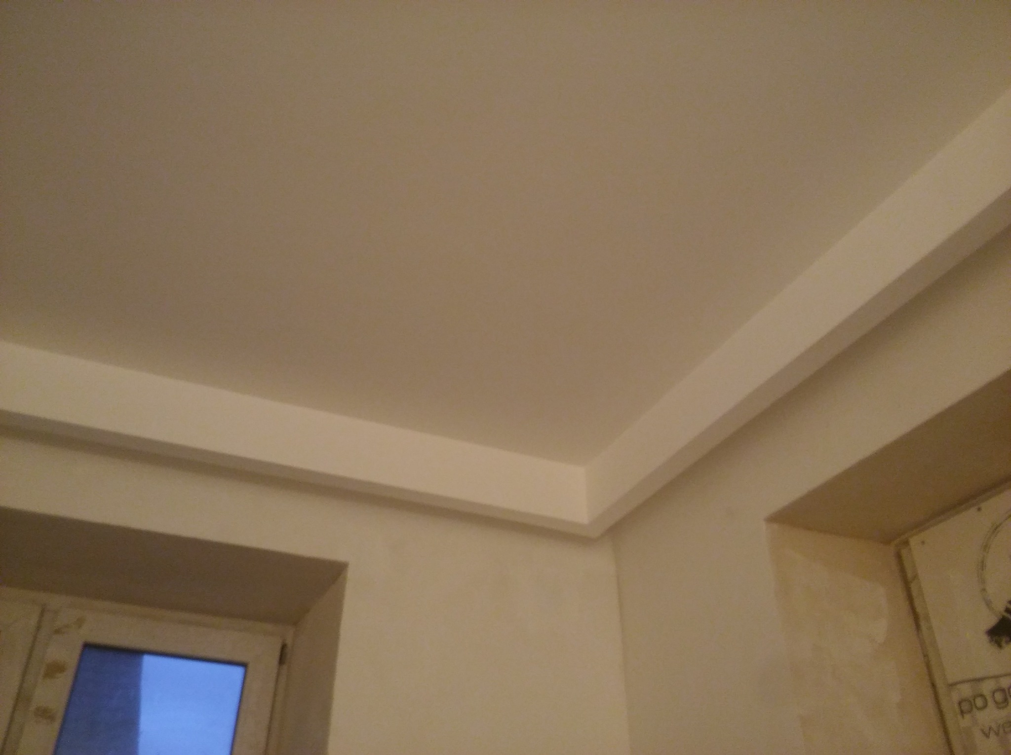 Wykończony narożnik sufitu z listwą dekoracyjną w jasnym pomieszczeniu, widoczny fragment okna i ściany, oświetlenie górne, lekko niedoskonałe połączenie ściany z sufitem.