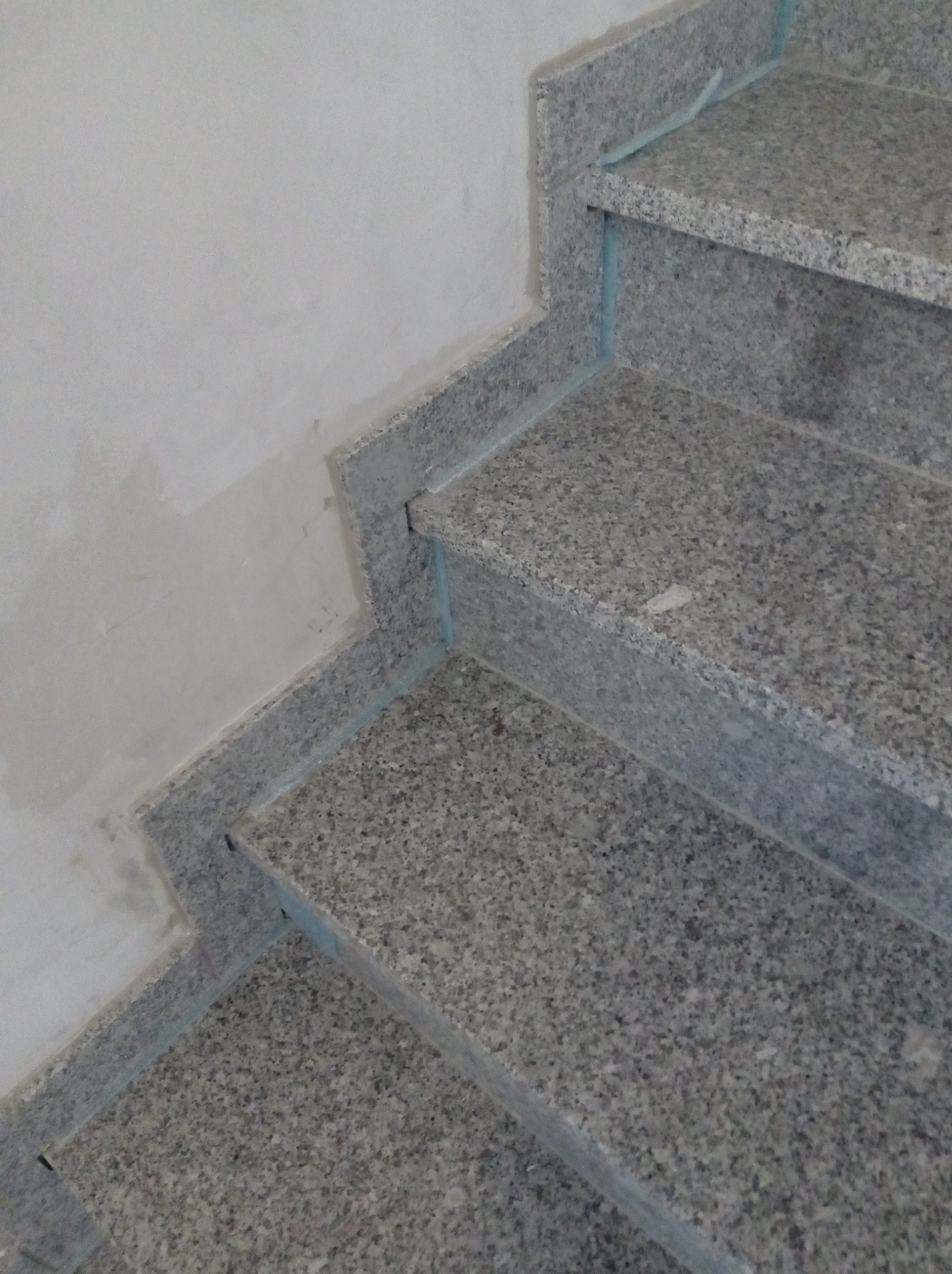 Wyłożone szarym kamieniem schody wewnętrzne, z widocznym fragmentem ściany w trakcie wykańczania i niebieską taśmą zabezpieczającą krawędzie.