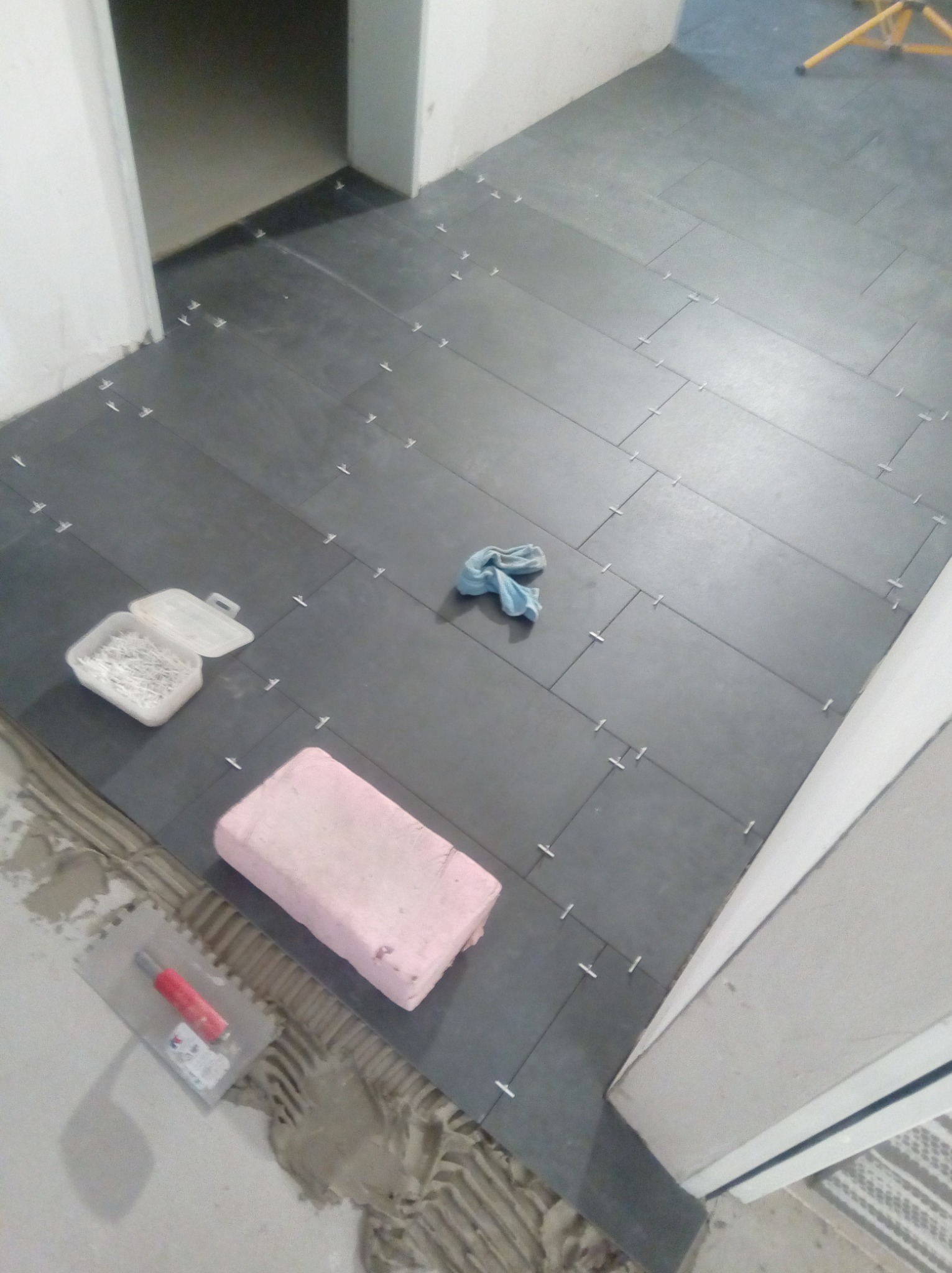 Proces układania ciemnych płytek podłogowych z użyciem krzyżyków dystansowych, widoczna paca z czerwoną rączką, pojemnik z klinami i gąbka. Świeżo nałożony klej pod płytkami.