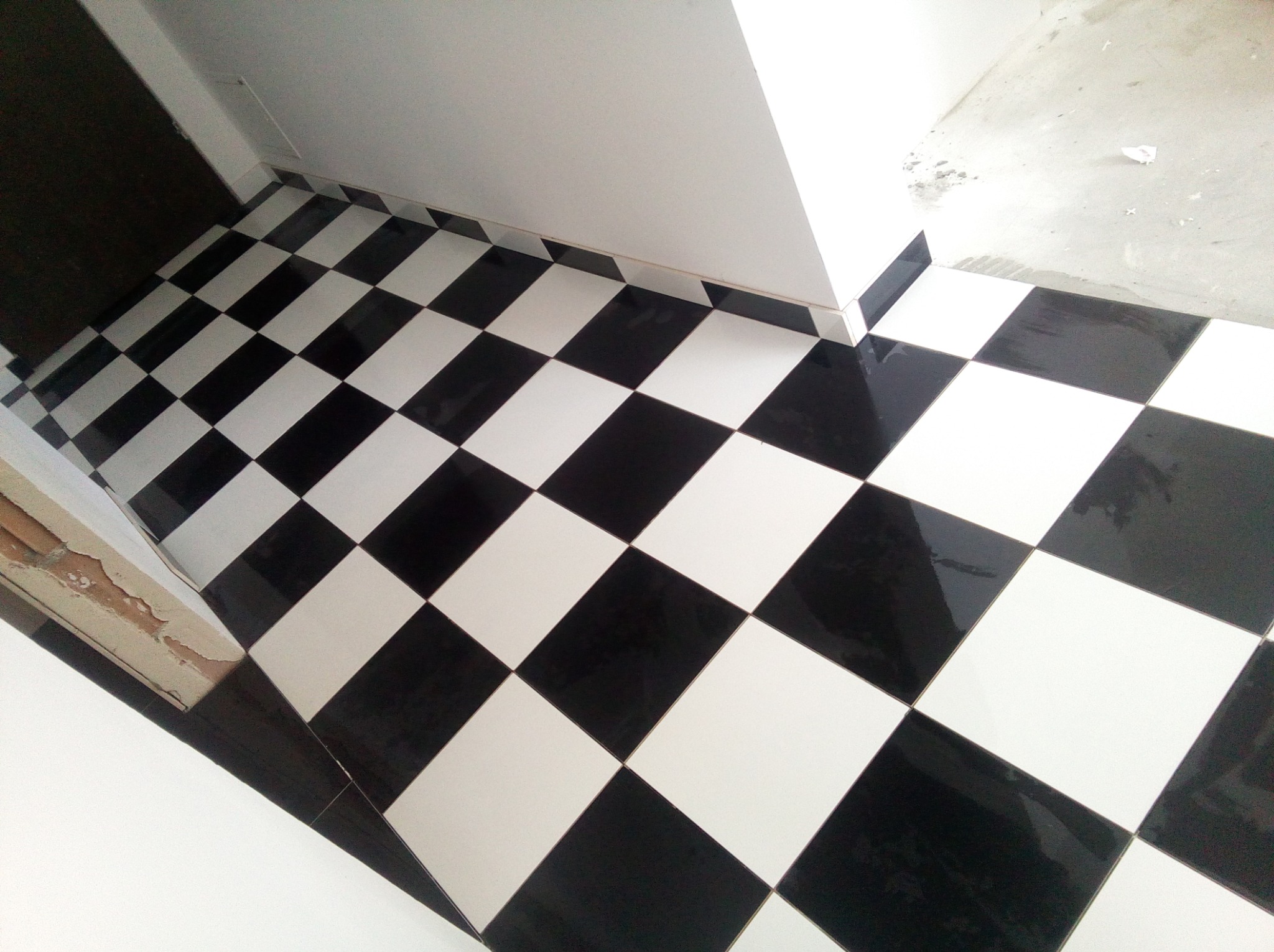 Ułożona pod kątem podłoga w czarno-białą szachownicę z błyszczących płytek, widoczny fragment niedokończonej ściany i futryna drzwi.