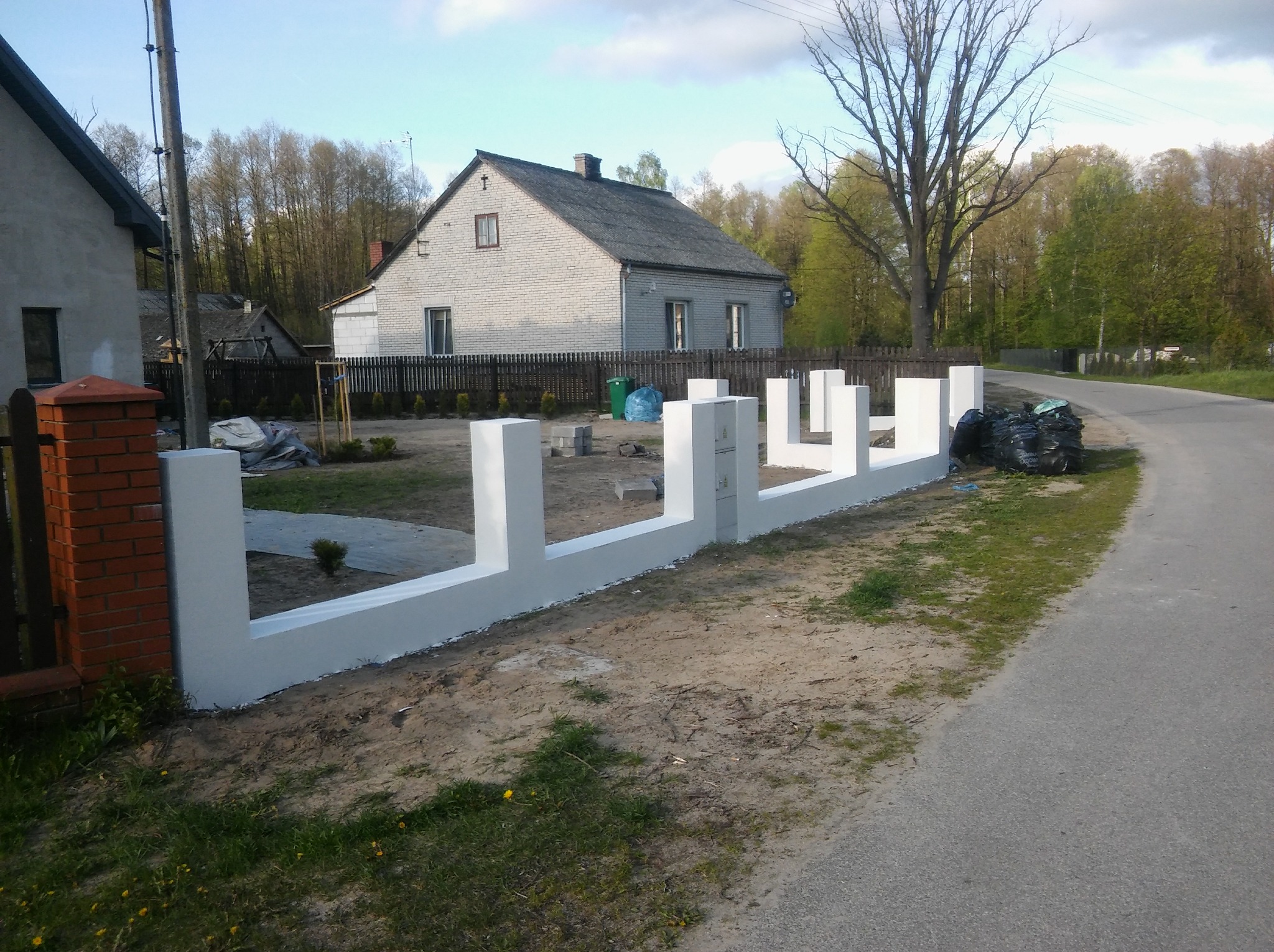 Nowy, biały, betonowy płot w trakcie budowy w Ostrołęce, widoczne słupki i segmenty, w tle dom jednorodzinny i worki z odpadami budowlanymi.