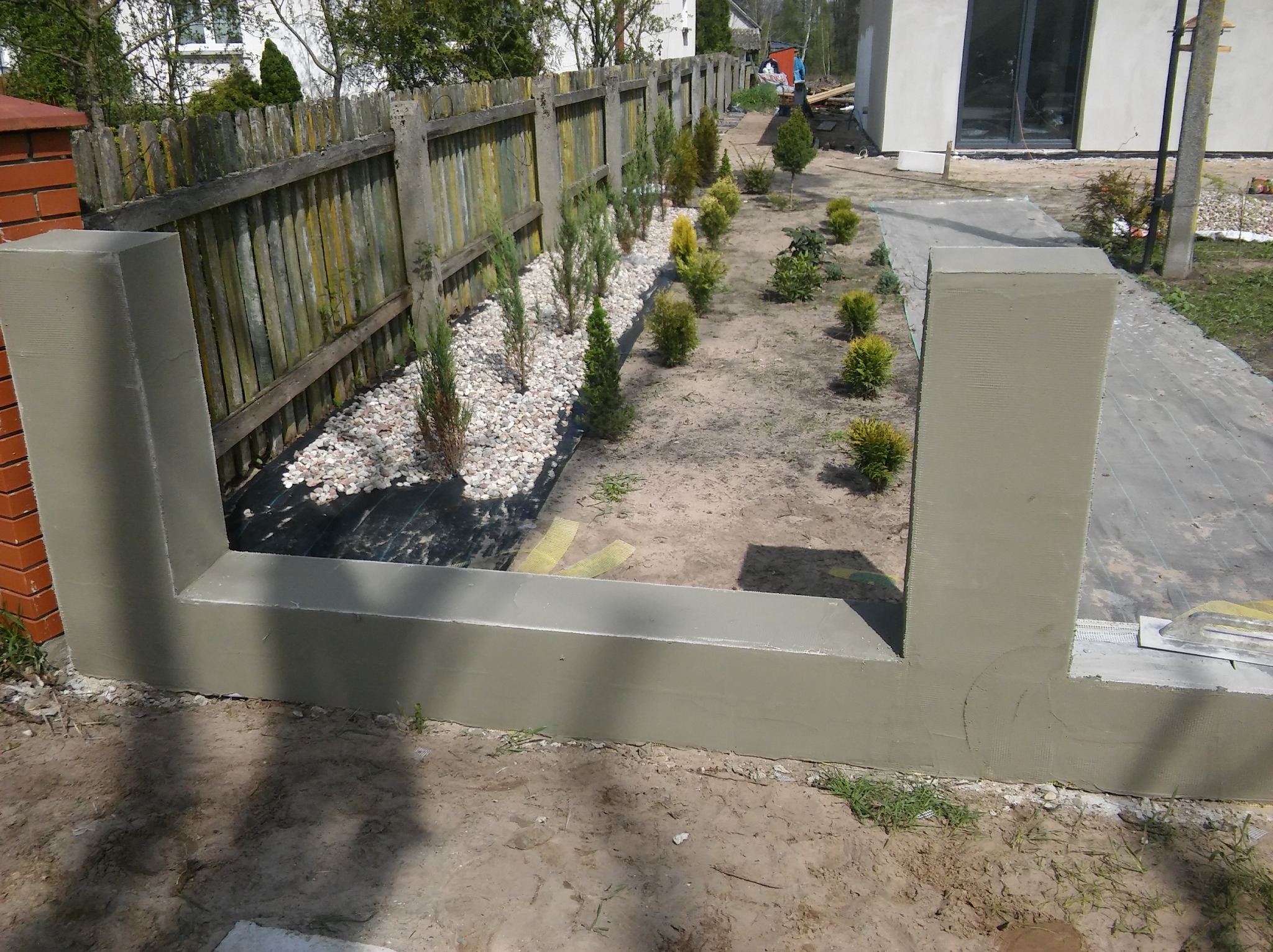 Świeżo otynkowany, niski murek z dwoma filarami, otaczający rabatę z młodymi tujami i kamykami, widoczny stary, drewniany płot w tle.