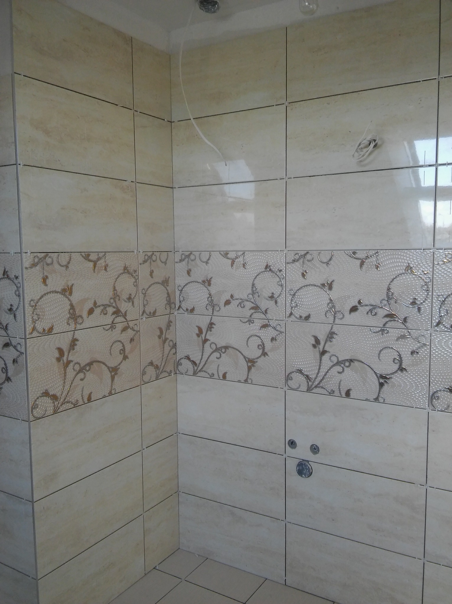 Fragment łazienki z beżowymi płytkami, w tym dekoracyjnymi z motywem roślinnym, widoczne krzyżyki dystansowe, przygotowane miejsce na baterię