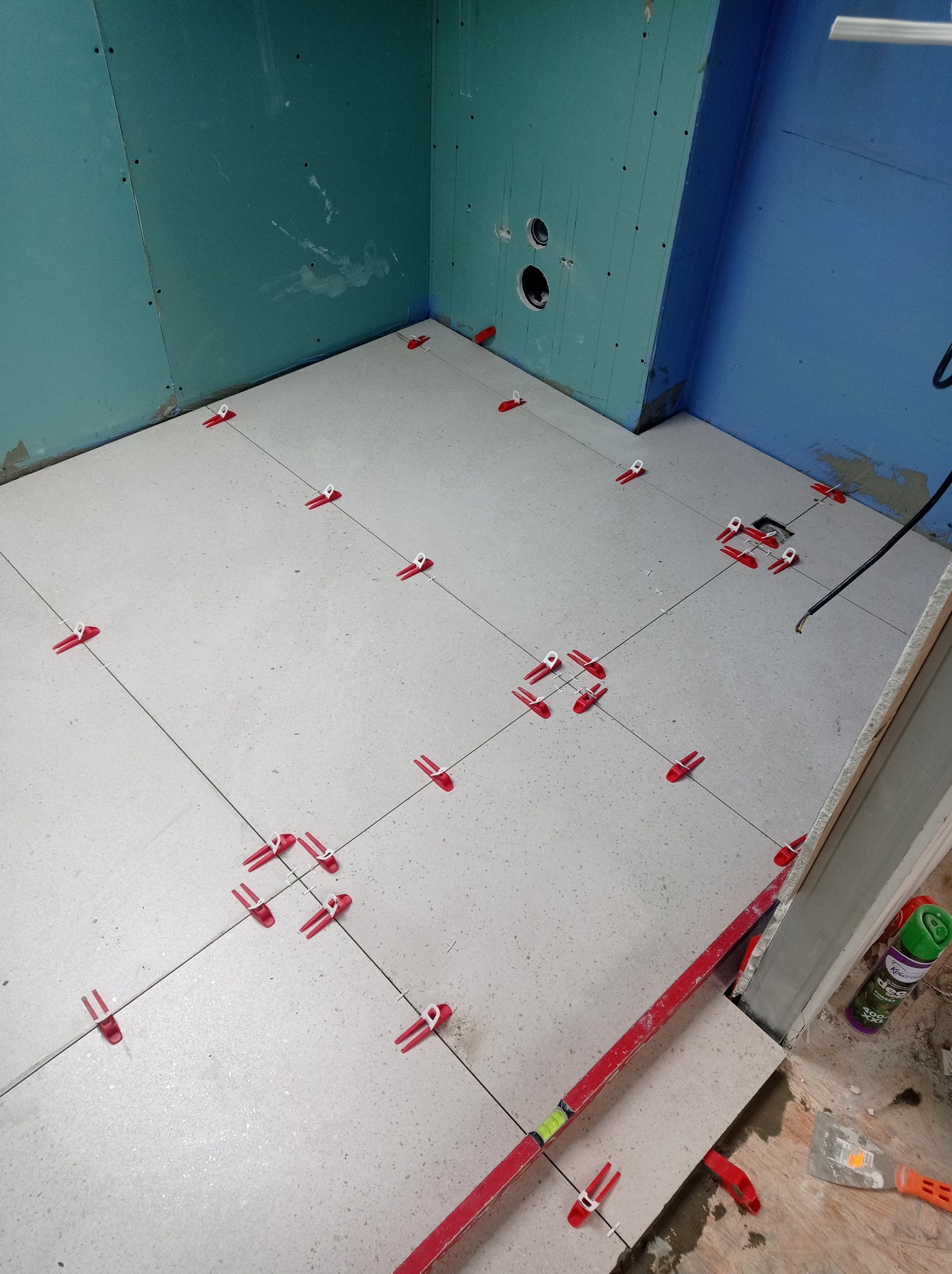 Układanie jasnych płytek podłogowych w łazience z użyciem systemu poziomowania z czerwonymi klipsami, widoczne ściany przygotowane pod wykończenie i poziomica w rogu pomieszczenia.