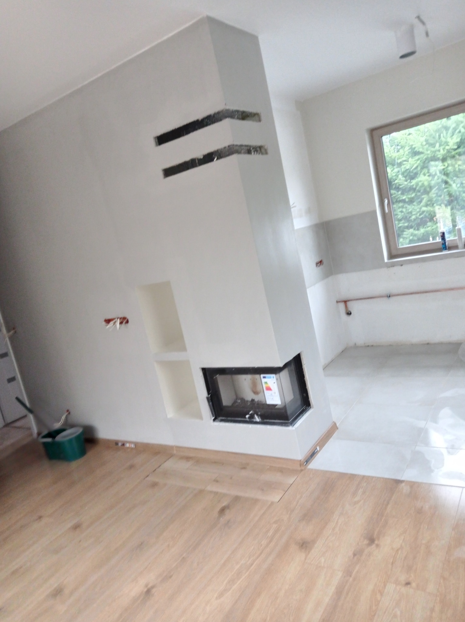 Wnętrze w trakcie wykańczania: obudowa kominka z wnękami, częściowo położona podłoga z paneli, widoczne instalacje elektryczne i hydrauliczne, oraz fragment łazienki z szarą glazurą.