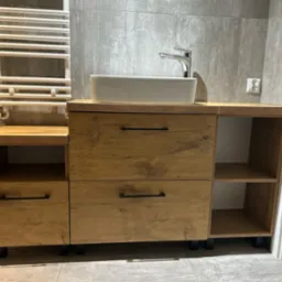 Nowoczesna szafka łazienkowa z drewnianym frontem, białą umywalką nablatową i chromowaną baterią, na tle szarych płytek i białego grzejnika drabinkowego.