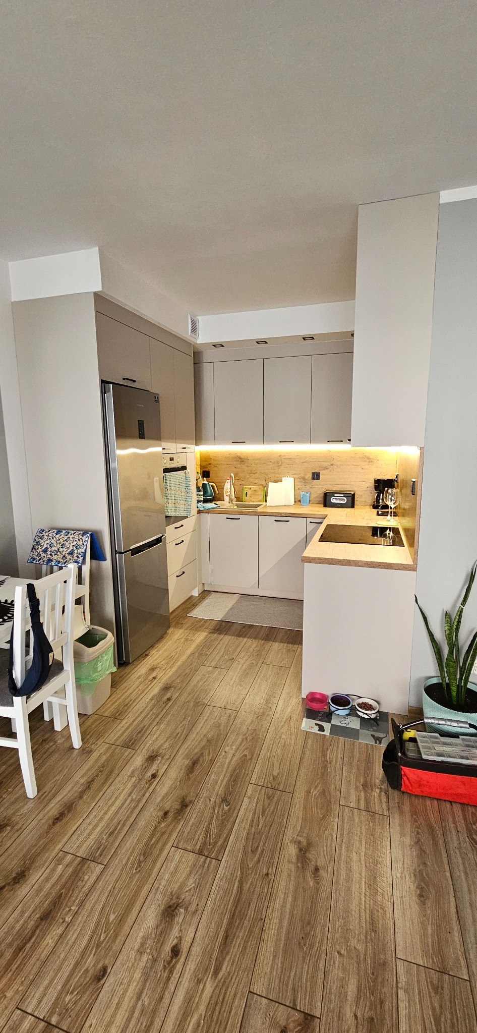 Jasna kuchnia z drewnianym blatem i oświetleniem LED pod szafkami, widoczny fragment jadalni i akcesoria dla zwierząt.