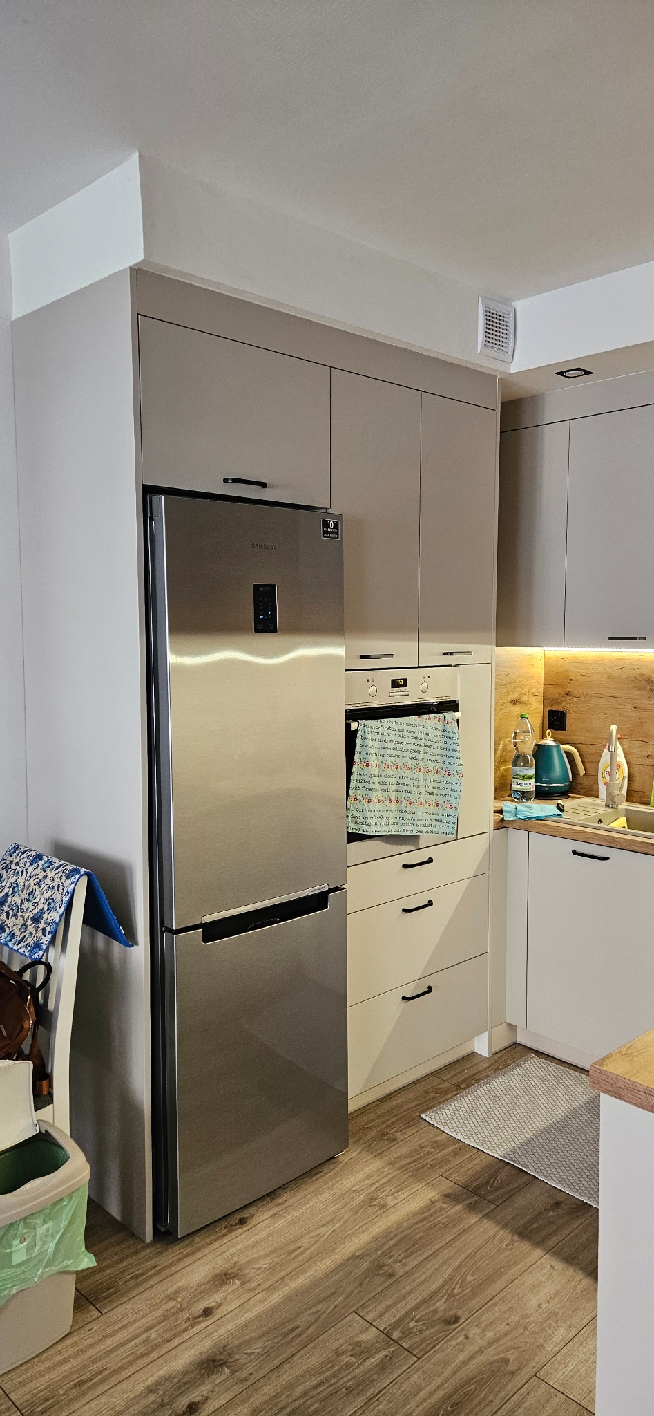 Nowoczesna kuchnia z zabudową w odcieniach szarości, z lodówką Samsung z wyświetlaczem, piekarnikiem z ręcznikiem z napisem, drewnianym blatem i oświetleniem LED podszafkowym.