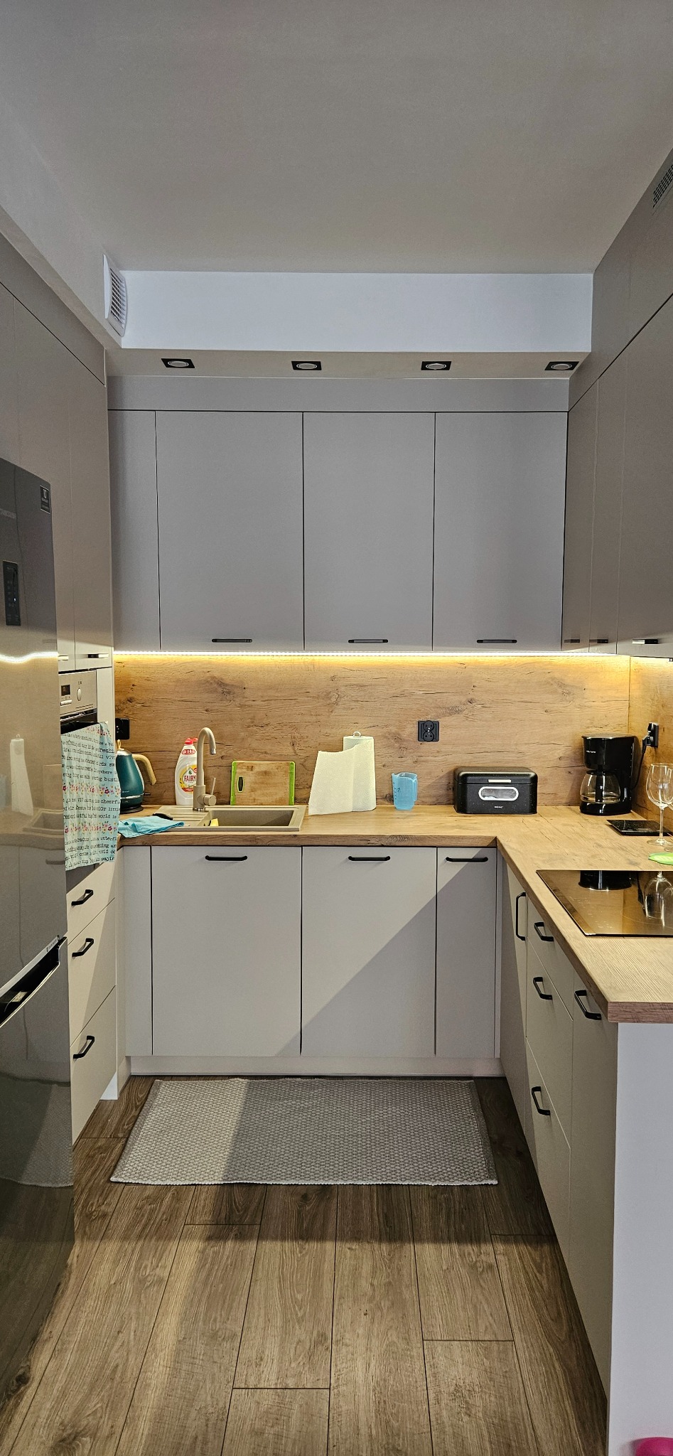 Szara kuchnia z drewnianym blatem i oświetleniem LED pod szafkami wiszącymi, widok na zlew, ekspres do kawy i toster na blacie.