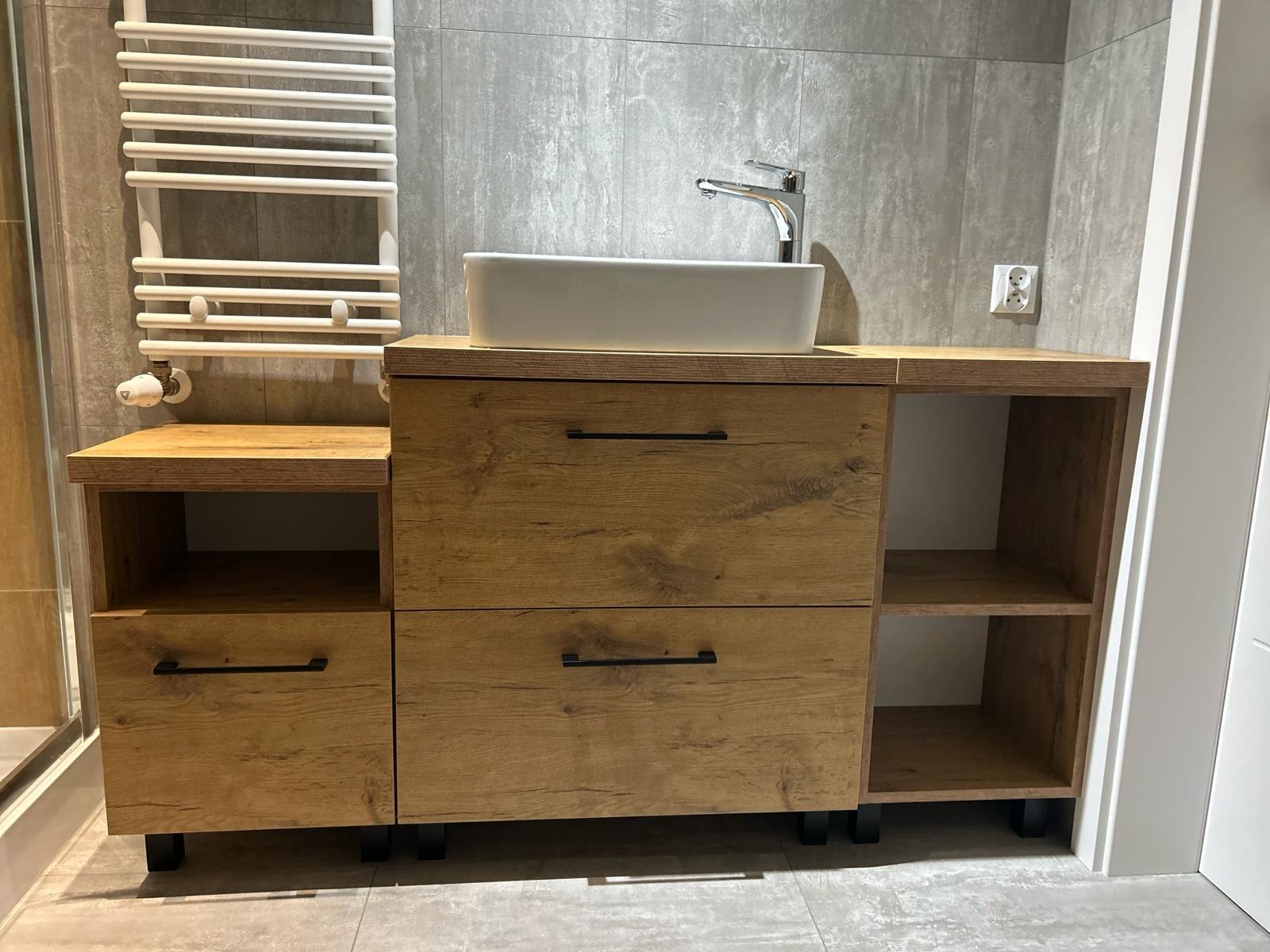 Nowoczesna szafka łazienkowa z drewnianym frontem, białą umywalką nablatową i chromowaną baterią, na tle szarych płytek i białego grzejnika drabinkowego.