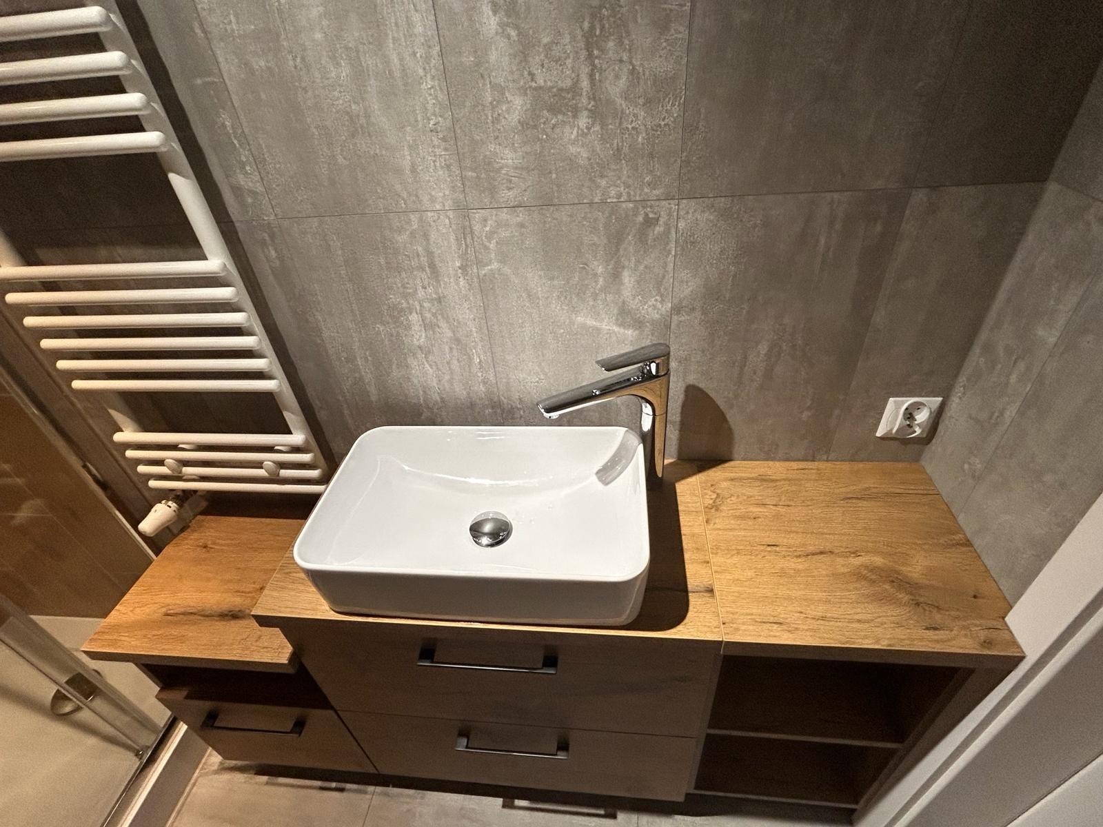 Nowoczesna łazienka z umywalką nablatową o kwadratowym kształcie, chromowaną baterią i szafką z drewnianym blatem. Na ścianie szare płytki imitujące beton oraz biały grzejnik drabinkowy.