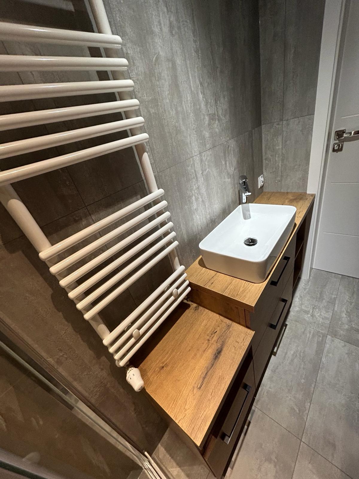 Nowoczesna łazienka z umywalką nablatową na drewnianej szafce z szufladami i białym grzejnikiem drabinkowym na tle szarych płytek.