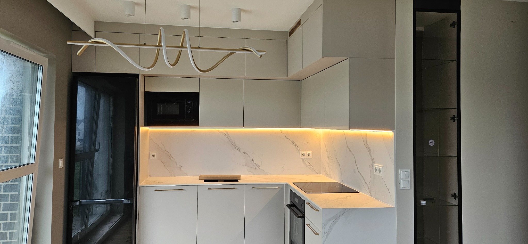 Nowoczesna kuchnia z szarymi frontami, marmurowym blatem i podświetleniem LED, widoczna czarna lodówka i witryna.