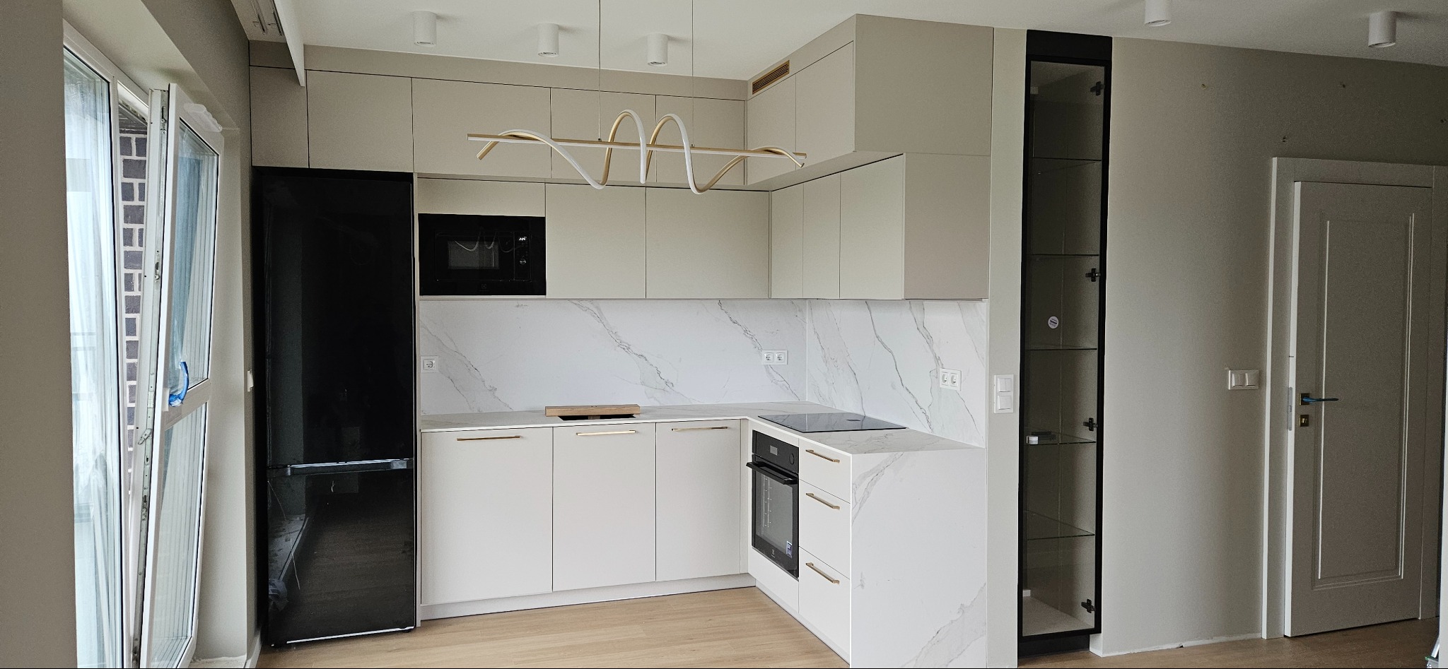 Jasna kuchnia z zabudową pod sufit, czarnym sprzętem AGD i marmurowym blatem, oświetlona nowoczesną lampą spiralną.