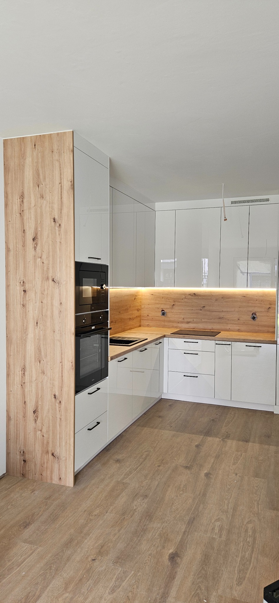 Nowoczesna kuchnia narożna z białymi, lakierowanymi frontami i drewnopodobnym panelem ściennym z oświetleniem LED, zabudowany piekarnik i płyta grzewcza.