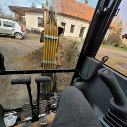 VERDI Karol Matera - Widok z kabiny koparki na wykop w ziemi, w tle dom z czerwoną dachówką i zaparkowany samochód.