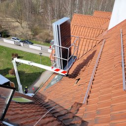 VERDI Karol Matera - Montaż paneli na dachu z użyciem podnośnika koszowego, widoczne szyny montażowe i pracownik w kamizelce odblaskowej na tle zieleni.