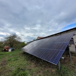VERDI Karol Matera - Panele słoneczne zamontowane na metalowej konstrukcji na trawiastym terenie, z budynkiem w tle i pochmurnym niebem.