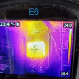 Wyświetlacz kamery termowizyjnej z zaznaczonym punktem pomiarowym na instalacji, widoczne temperatury w stopniach Celsjusza, oznaczenie E6 na obudowie urządzenia.