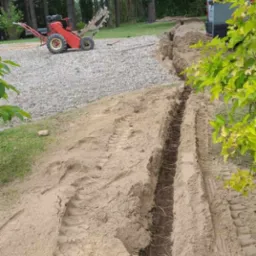 Wąski wykop w ziemi z widocznymi śladami opon, w tle czerwona glebogryzarka na kamienistym podłożu i częściowo widoczny samochód dostawczy.
