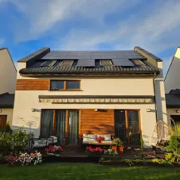 Nowoczesny dom jednorodzinny z panelami słonecznymi na dachu, widocznymi również oknami dachowymi, taras z meblami ogrodowymi i bujną zielenią w słoneczny dzień.