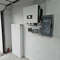 VERDI Karol Matera - Biały magazyn energii SOFAR obok inwertera SOFAR i dwóch skrzynek z bezpiecznikami na szarej ścianie garażu, widoczna gaśnica.