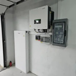 Biały magazyn energii SOFAR obok inwertera SOFAR i dwóch skrzynek z bezpiecznikami na szarej ścianie garażu, widoczna gaśnica.