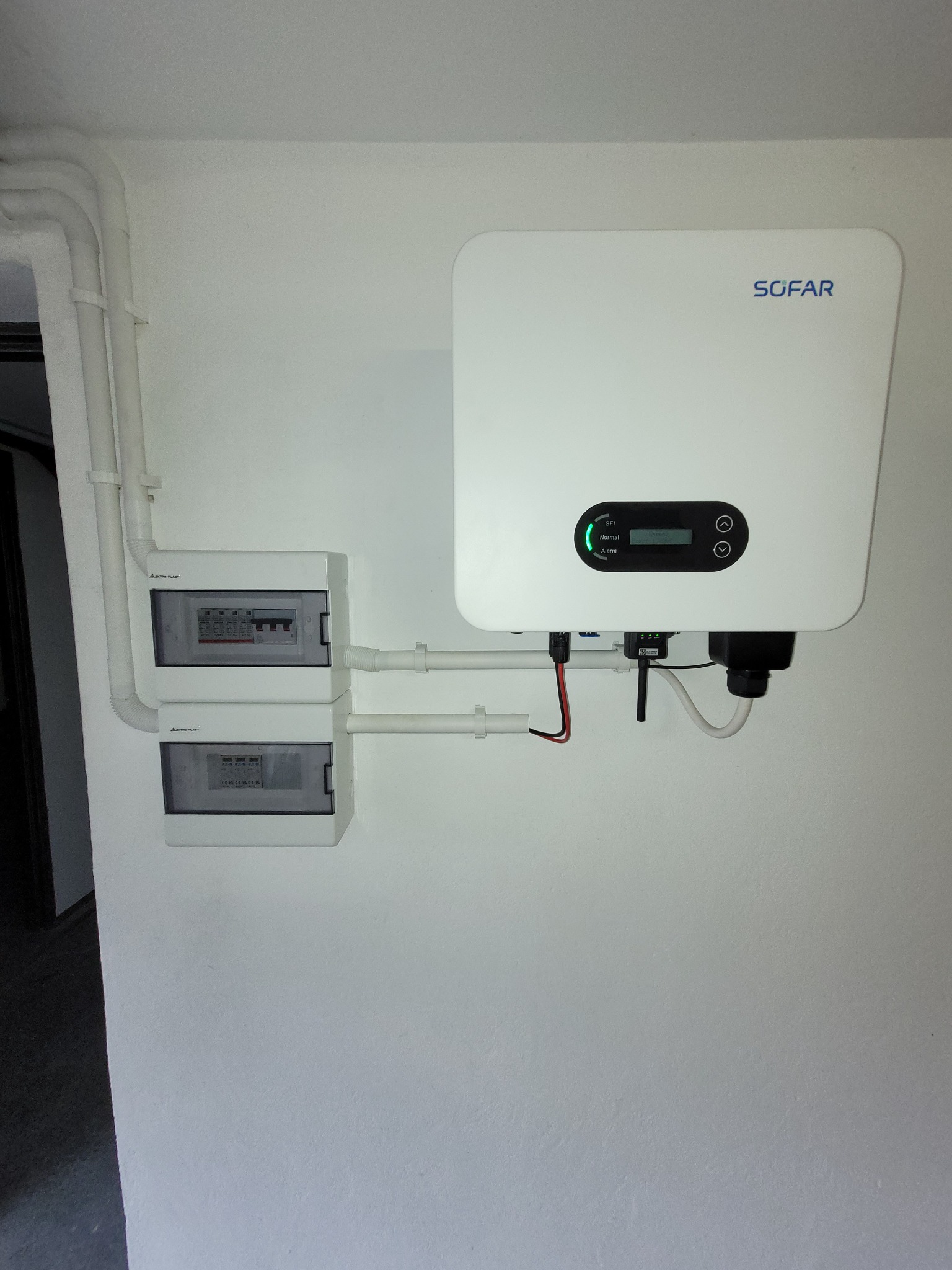 Biały inwerter SOFAR zamontowany na ścianie, połączony z dwiema podwójnymi skrzynkami bezpiecznikowymi i rurami PCV, widoczny zielony wskaźnik LED