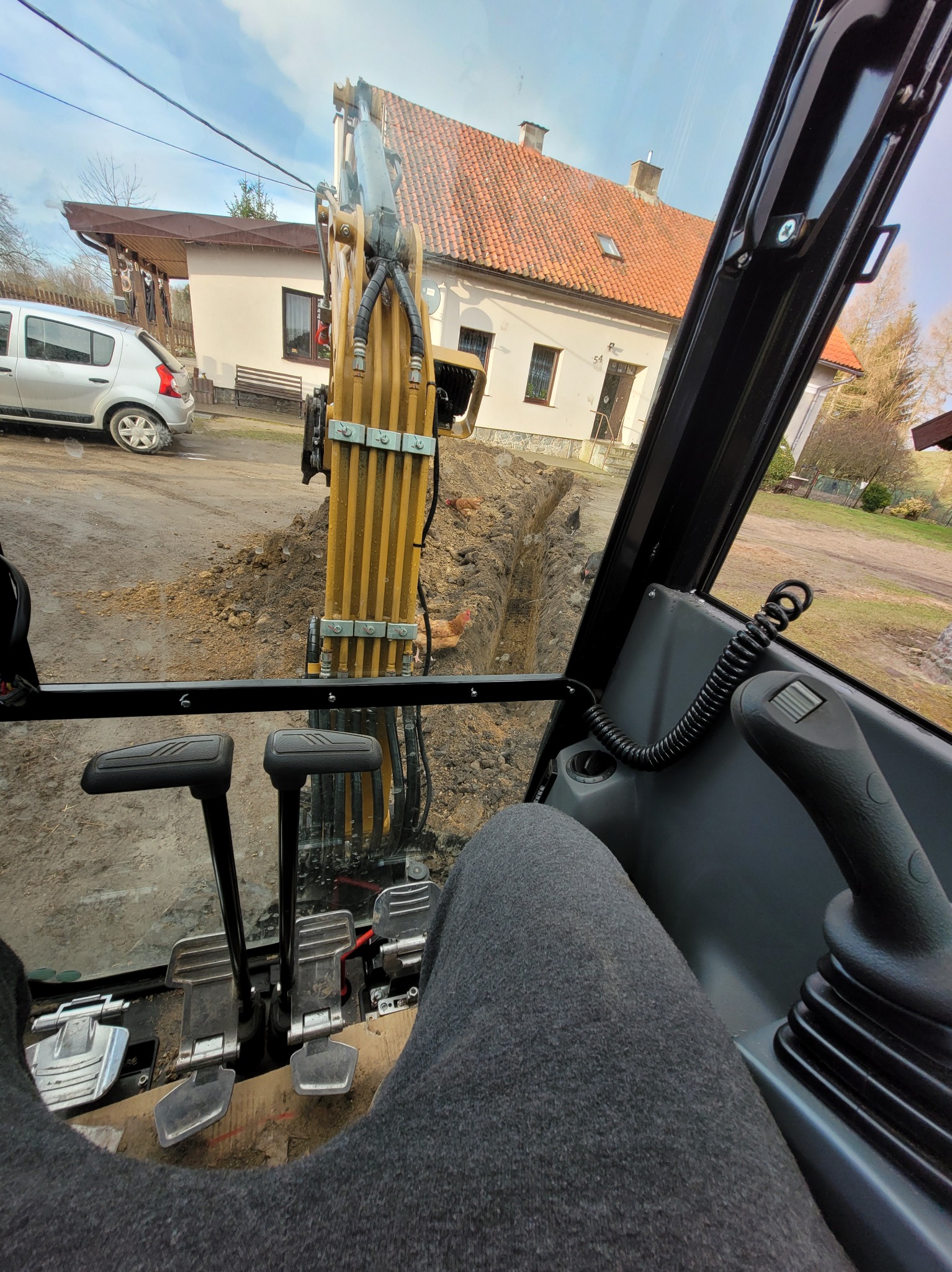 Widok z kabiny koparki na wykop w ziemi, w tle dom z czerwoną dachówką i zaparkowany samochód.