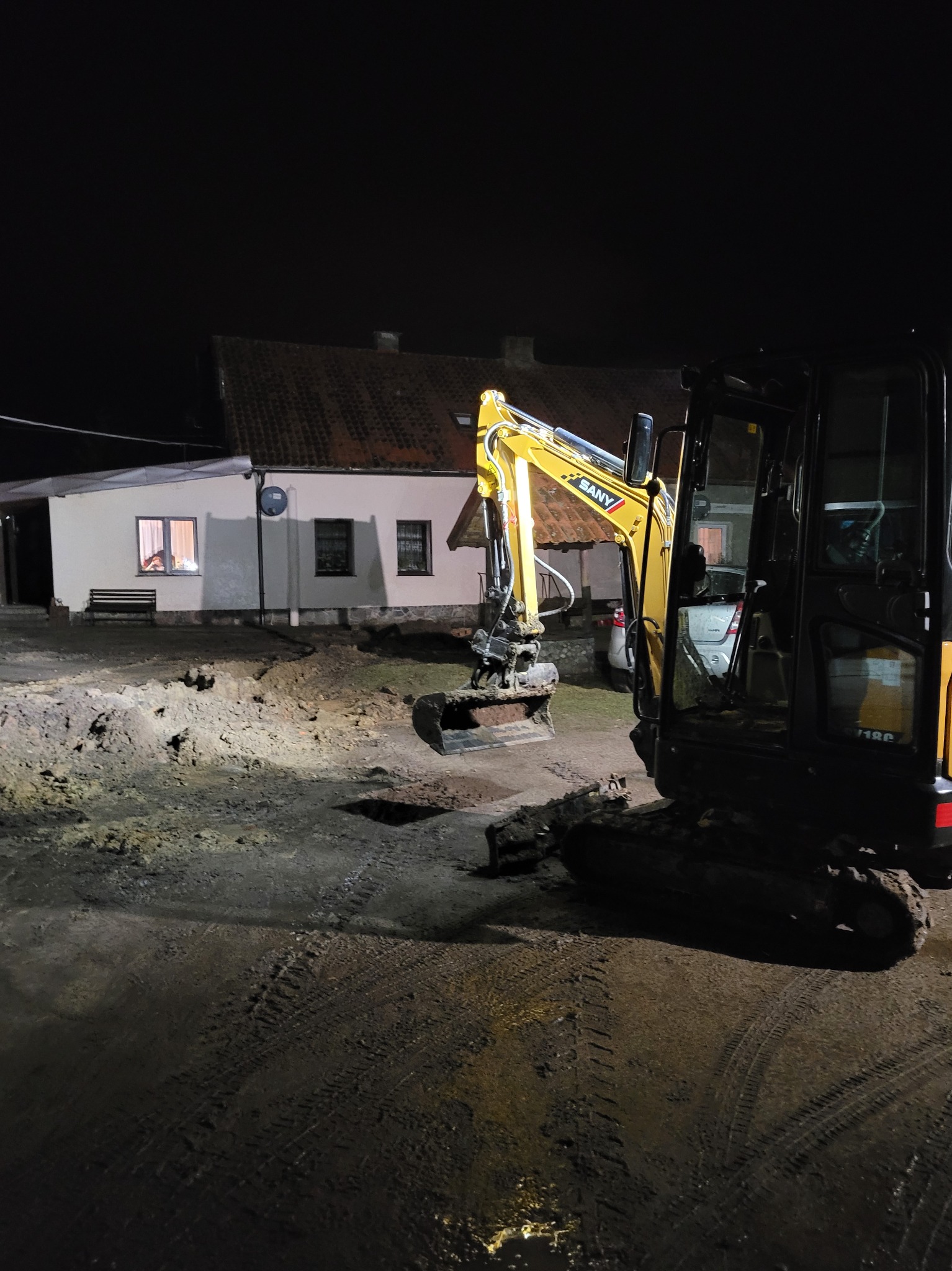 Żółto-czarna mini koparka SANY na placu budowy w nocy, oświetlona światłem z okien domu w tle.