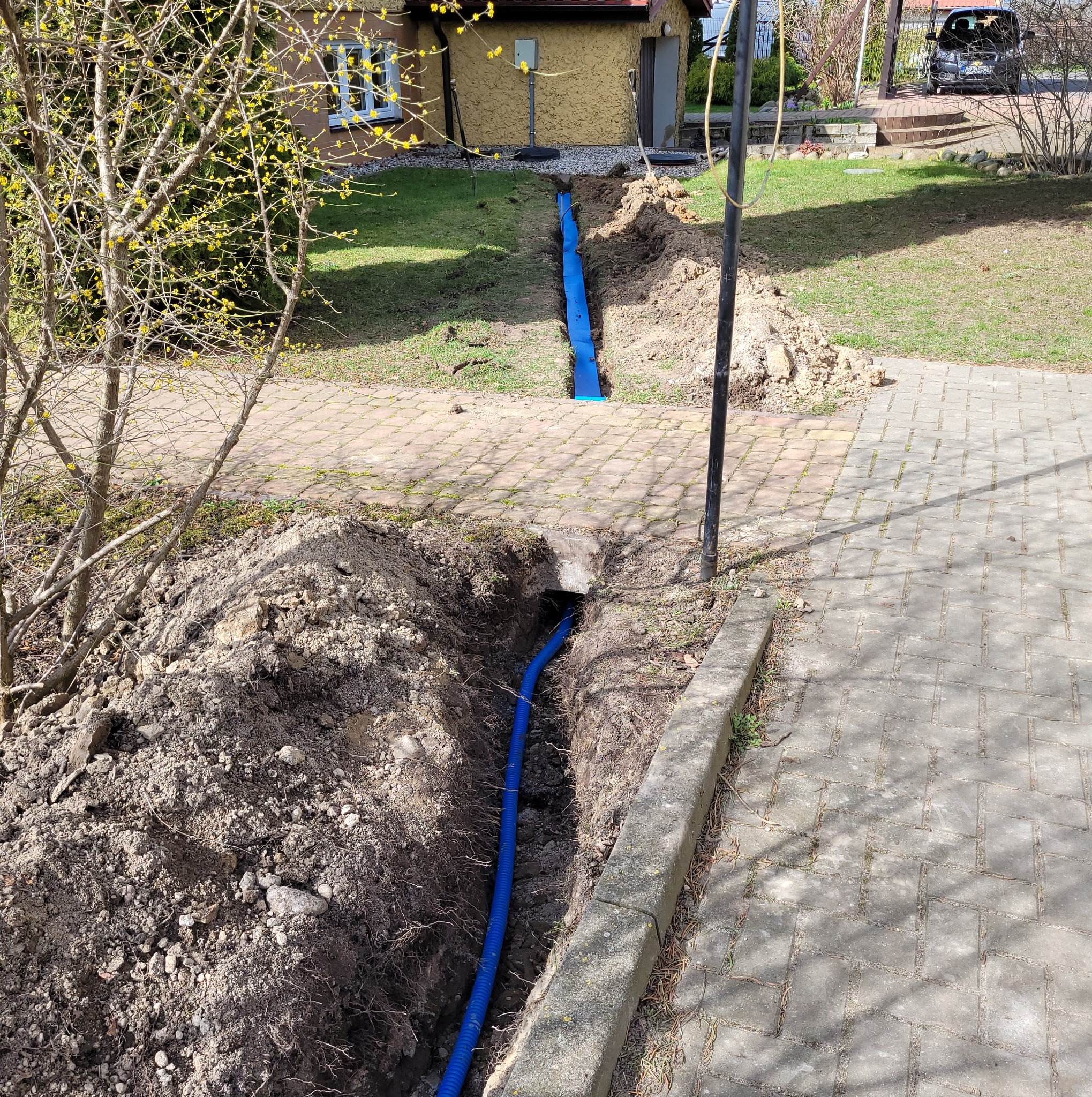 Wykonany wykop z ułożoną niebieską rurą karbowaną prowadzącą do budynku, ziemia usypana obok wykopu, kostka brukowa w tle