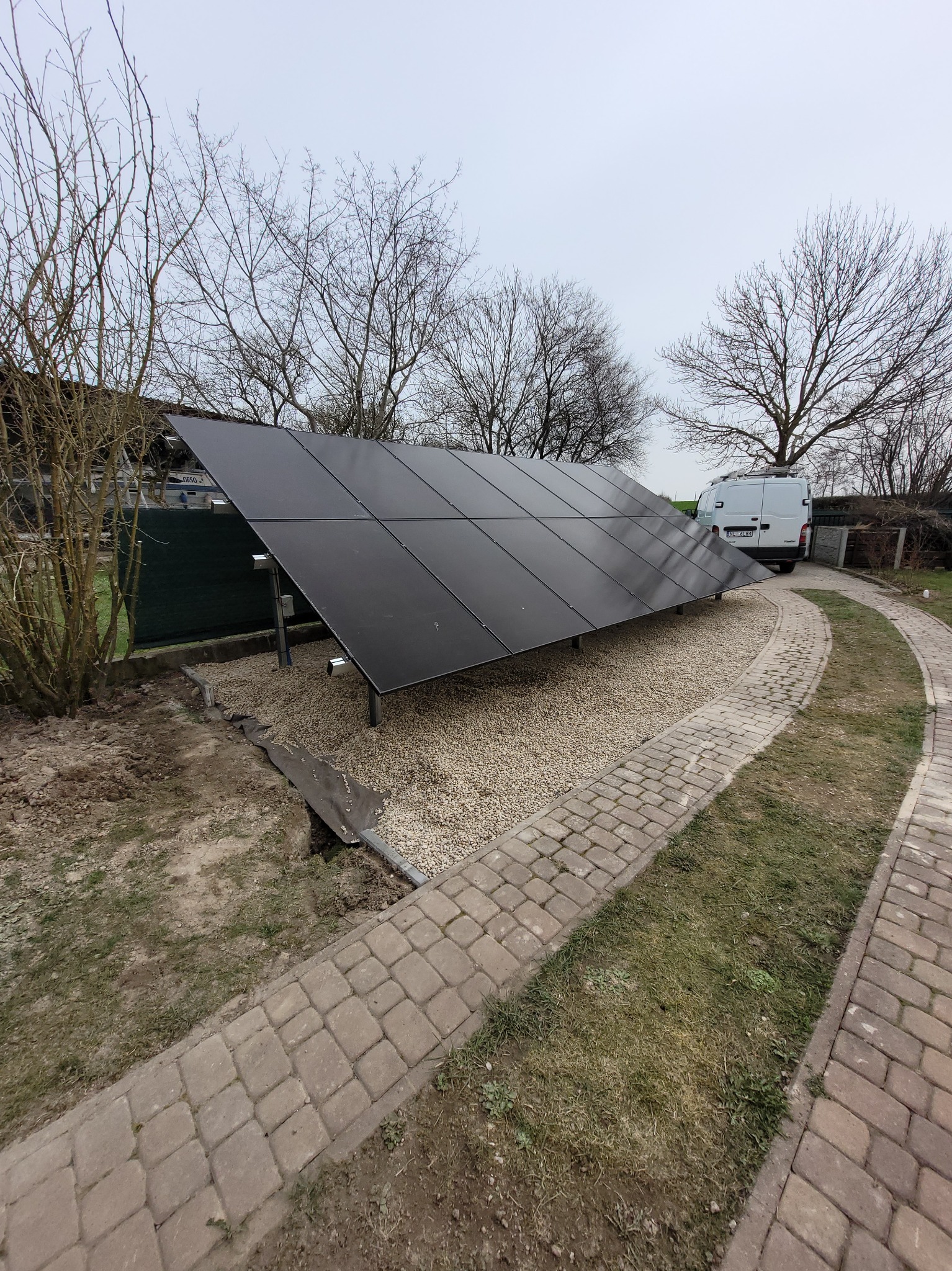 Czarny panel słoneczny ustawiony na żwirowym podłożu w ogrodzie, z widocznym białym vanem w tle i ścieżką z kostki brukowej.