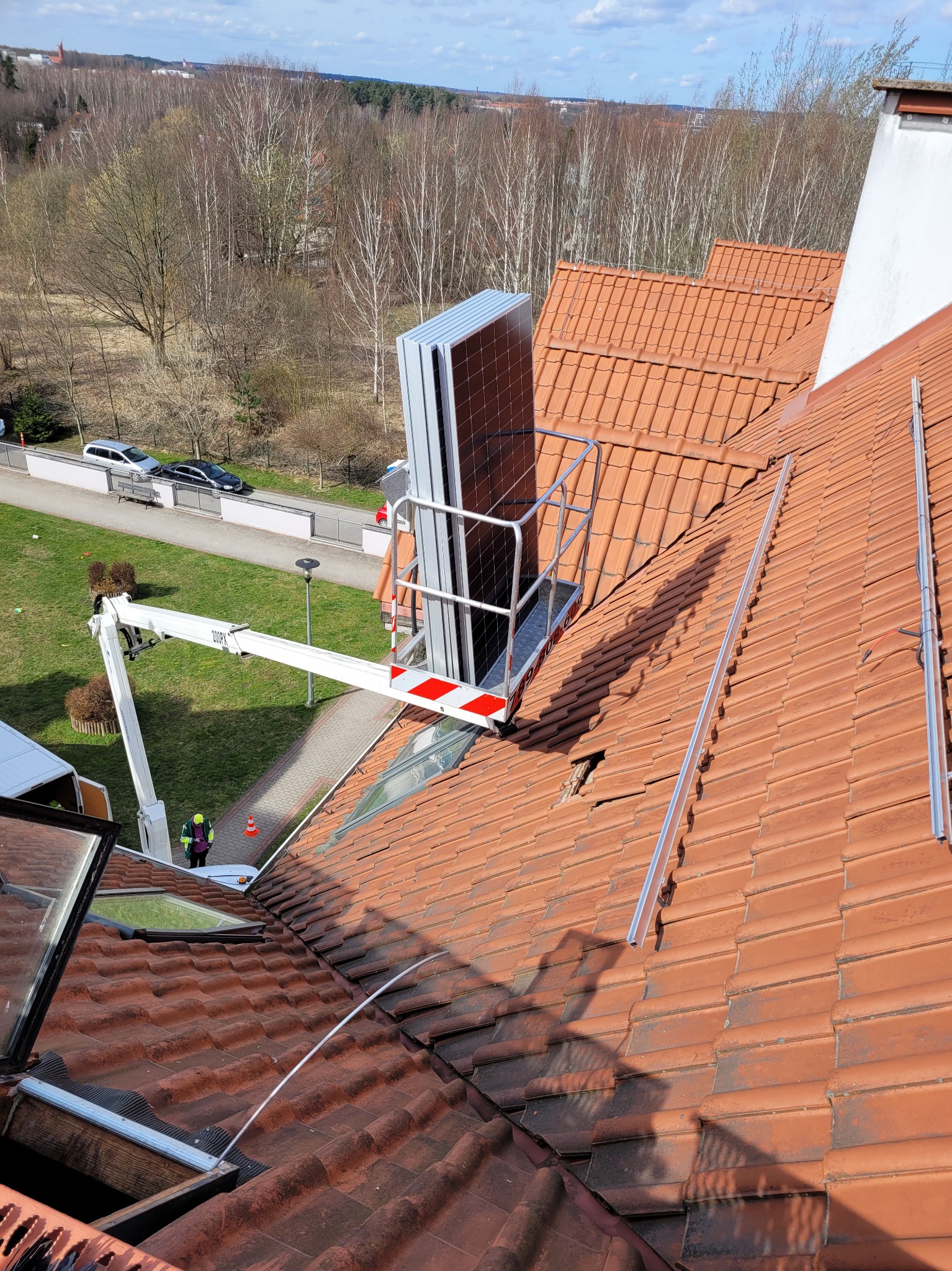 Montaż paneli na dachu z użyciem podnośnika koszowego, widoczne szyny montażowe i pracownik w kamizelce odblaskowej na tle zieleni.