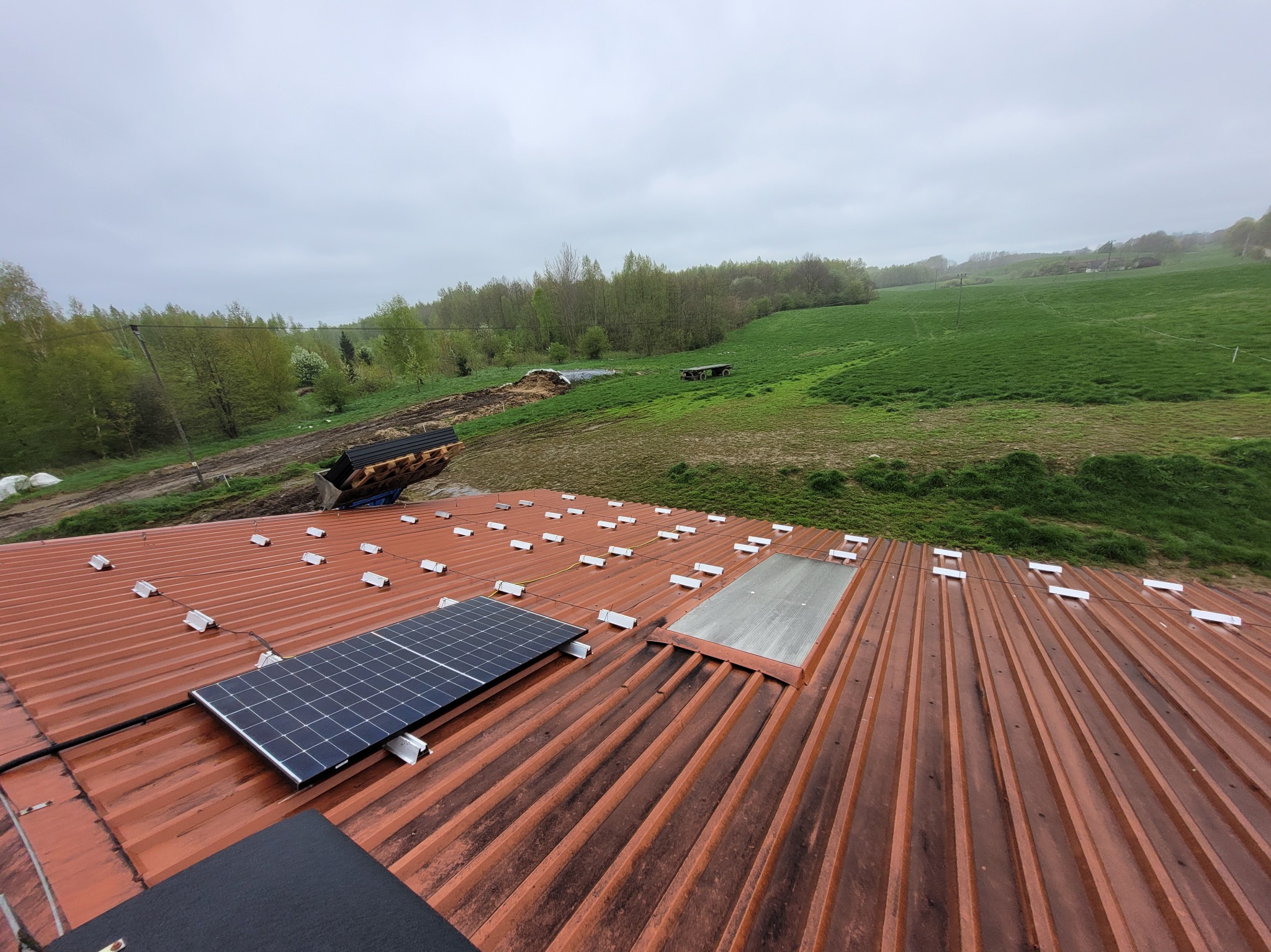 Montaż paneli słonecznych na dachu z blachy trapezowej, widok na krajobraz wiejski w pochmurny dzień.