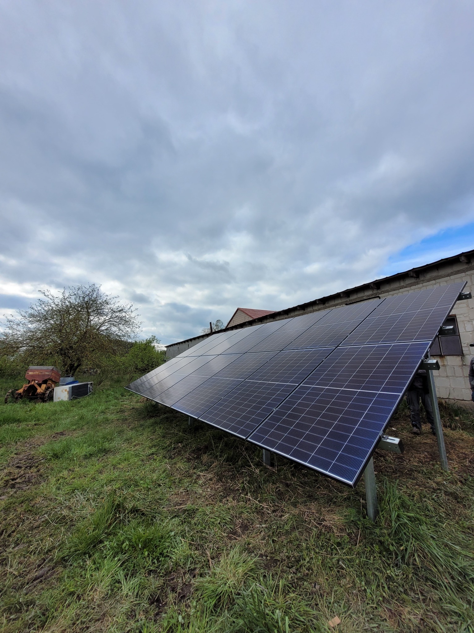 Panele słoneczne zamontowane na metalowej konstrukcji na trawiastym terenie, z budynkiem w tle i pochmurnym niebem.
