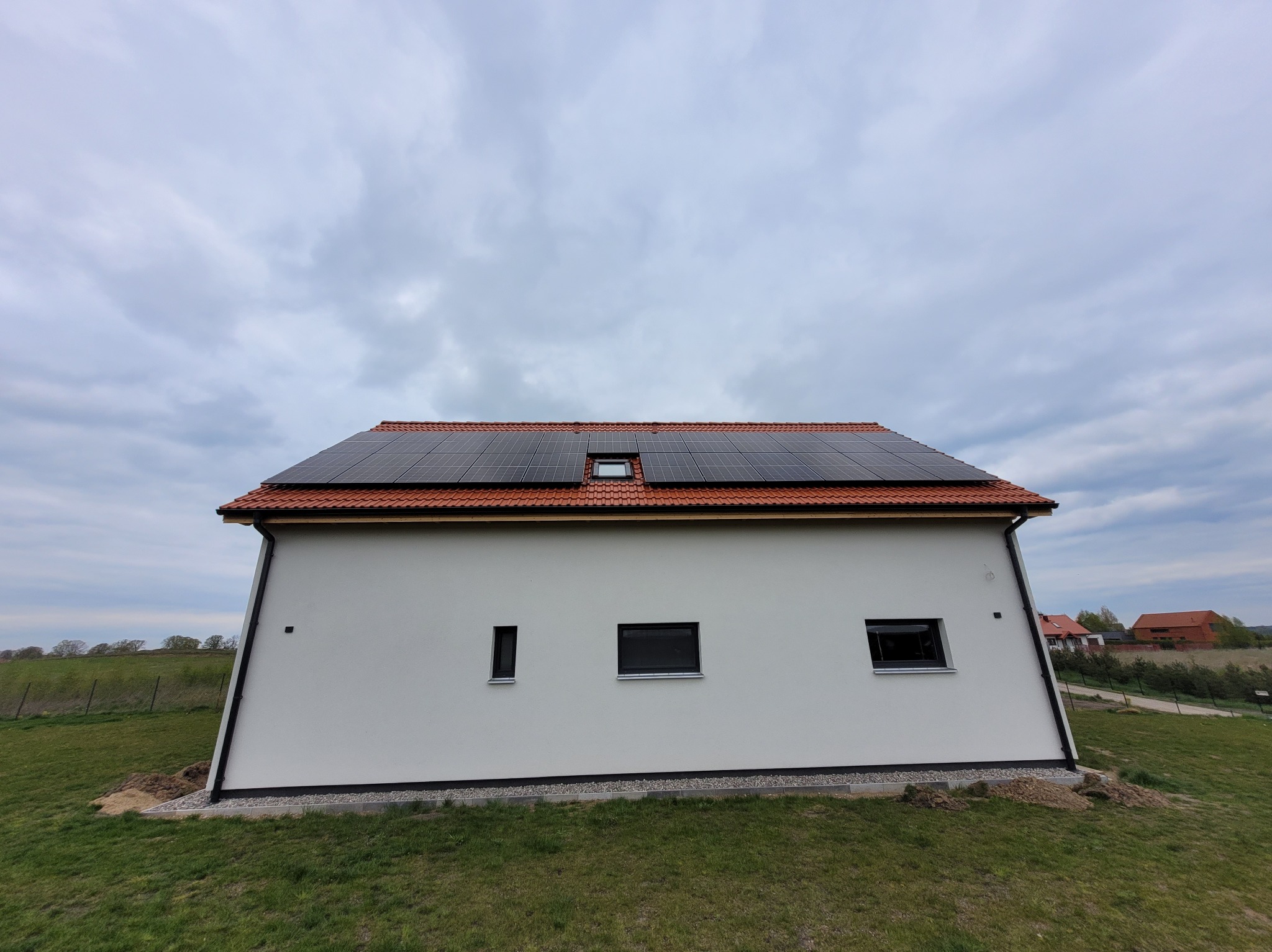 Boczny widok domu jednorodzinnego z zainstalowanymi panelami słonecznymi na dachu z czerwoną dachówką, małe okno dachowe, szara kostka brukowa przy ścianie, pochmurne niebo.