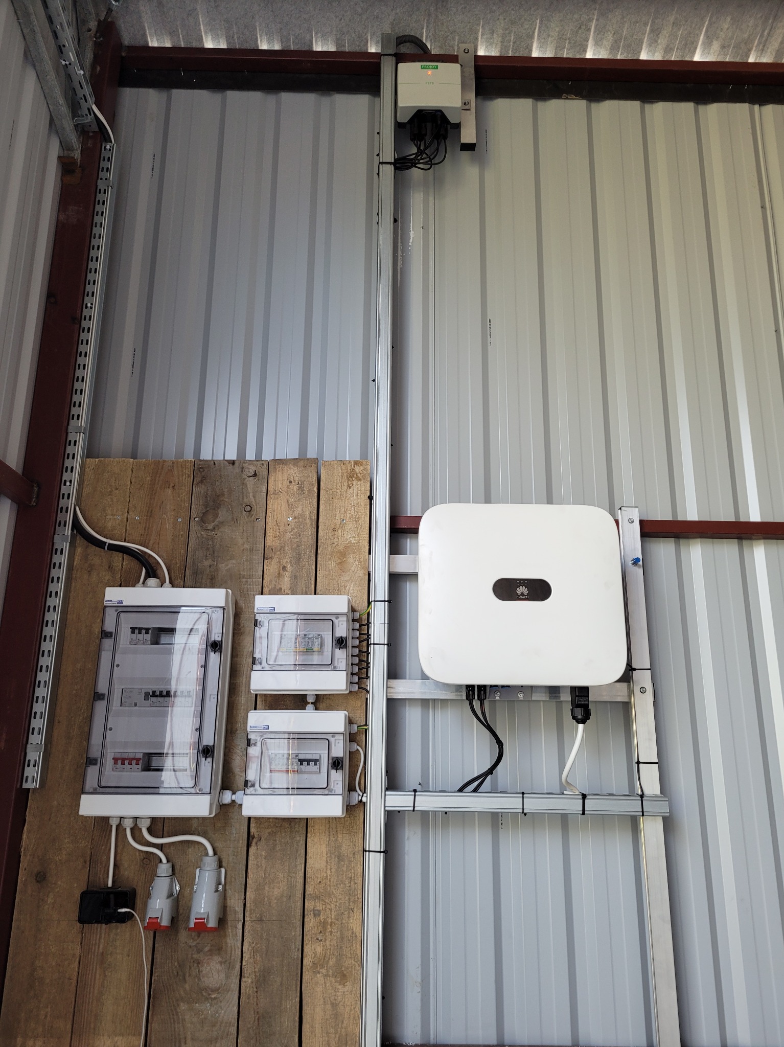 Biały inwerter Huawei zamontowany na metalowej konstrukcji na tle blachy trapezowej, obok drewnianej płyty z trzema rozdzielnicami elektrycznymi i gniazdami wtykowymi.