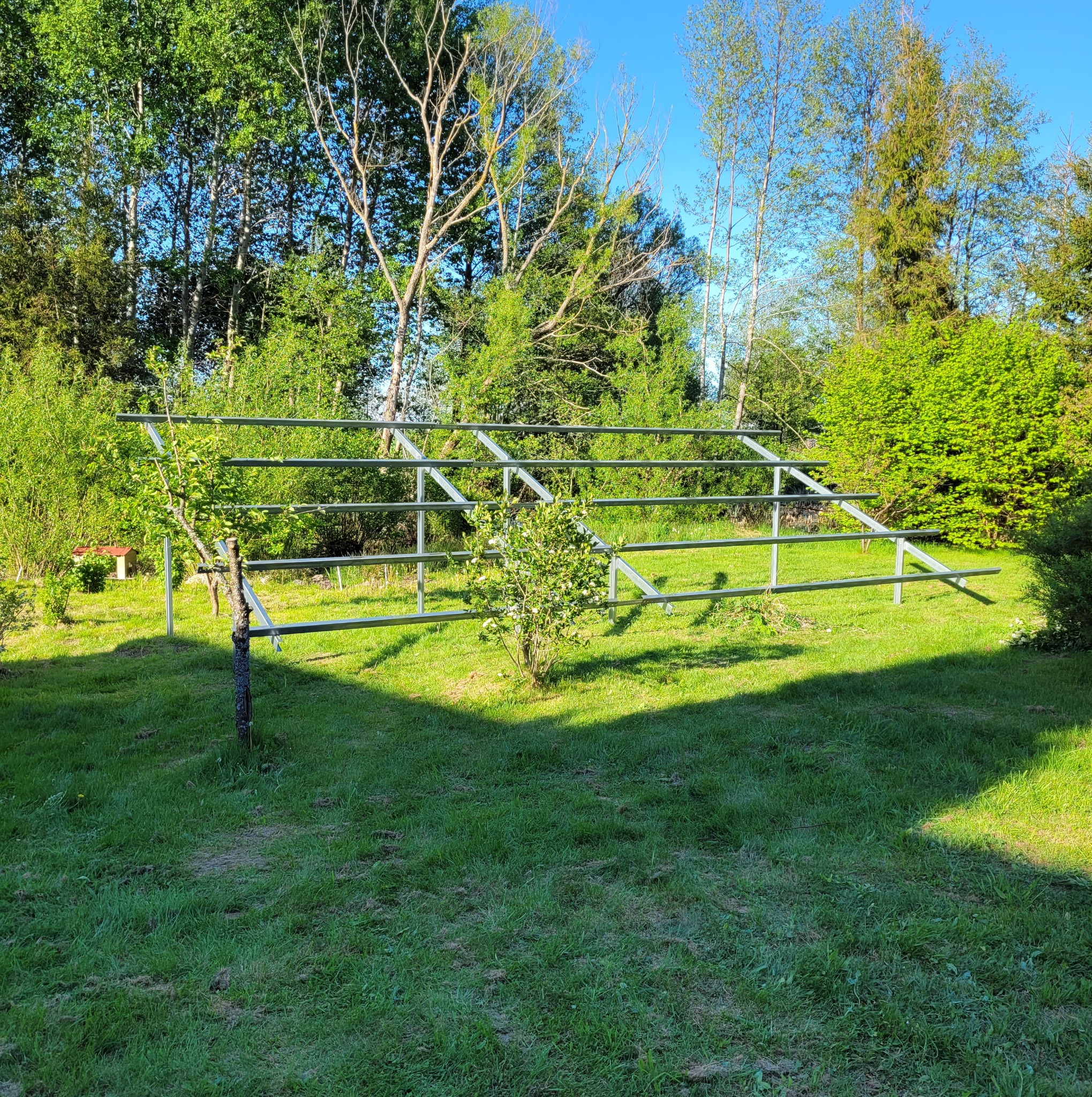 Stalowa konstrukcja wsporcza na trawniku, przygotowana pod montaż paneli, w tle drzewa i krzewy.